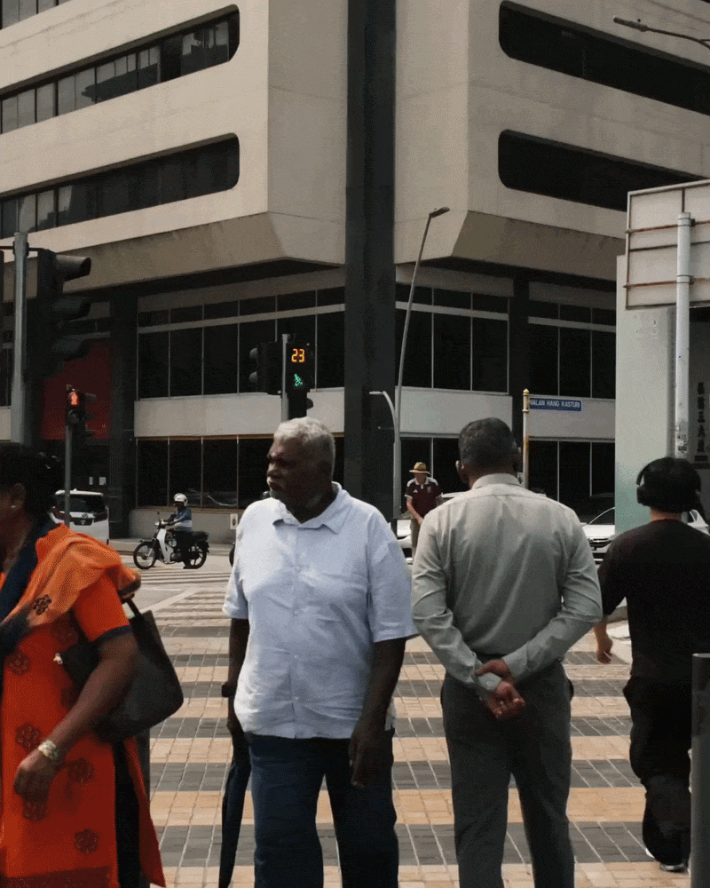 People move through a sunlit pedestrian crossing in Kuala Lumpur, with passing traffic and a modern concrete building in the background.
