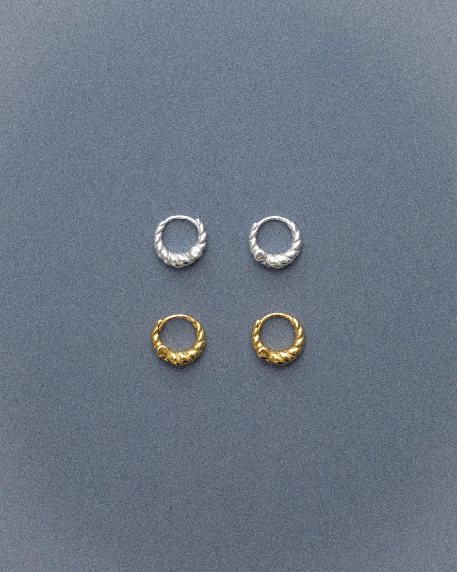 Two pairs of spiral hoop earrings, one in gold and one in silver, arranged in a delicate flatlay, their tiny forms resting on a quiet grey-blue background.