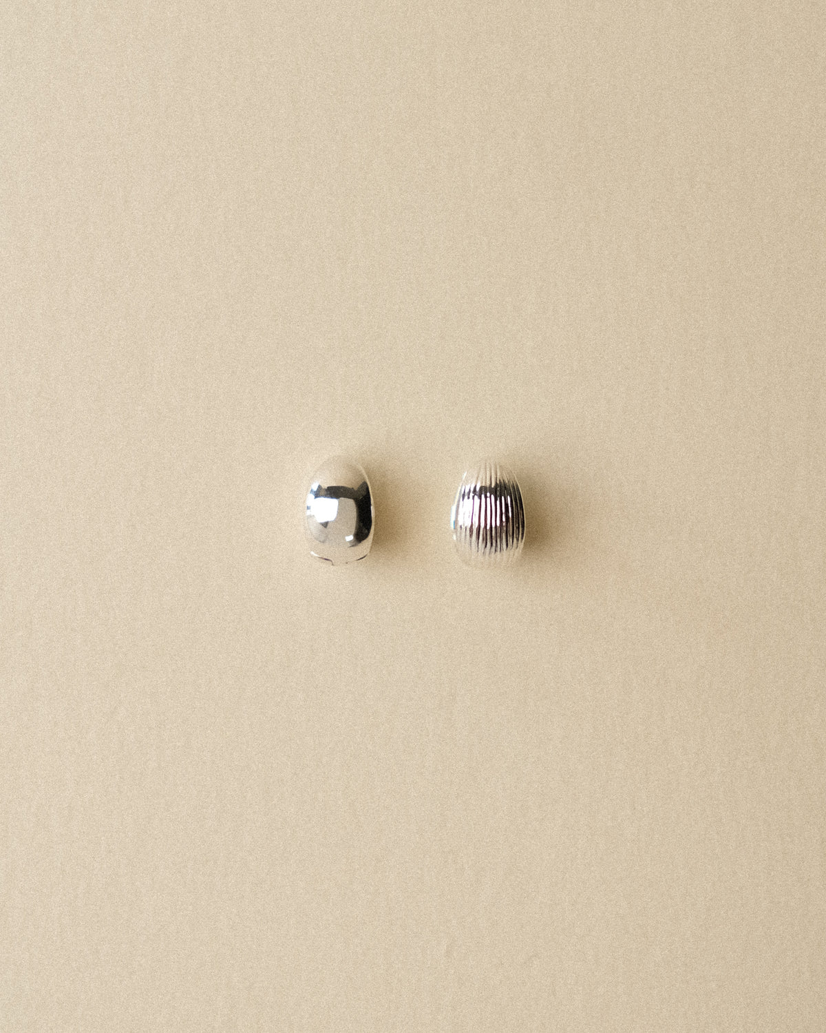 A pair of small silver-plated brass reversible hoop earrings, one smooth and one ribbed, displayed on a textured wabi-sabi warm background.