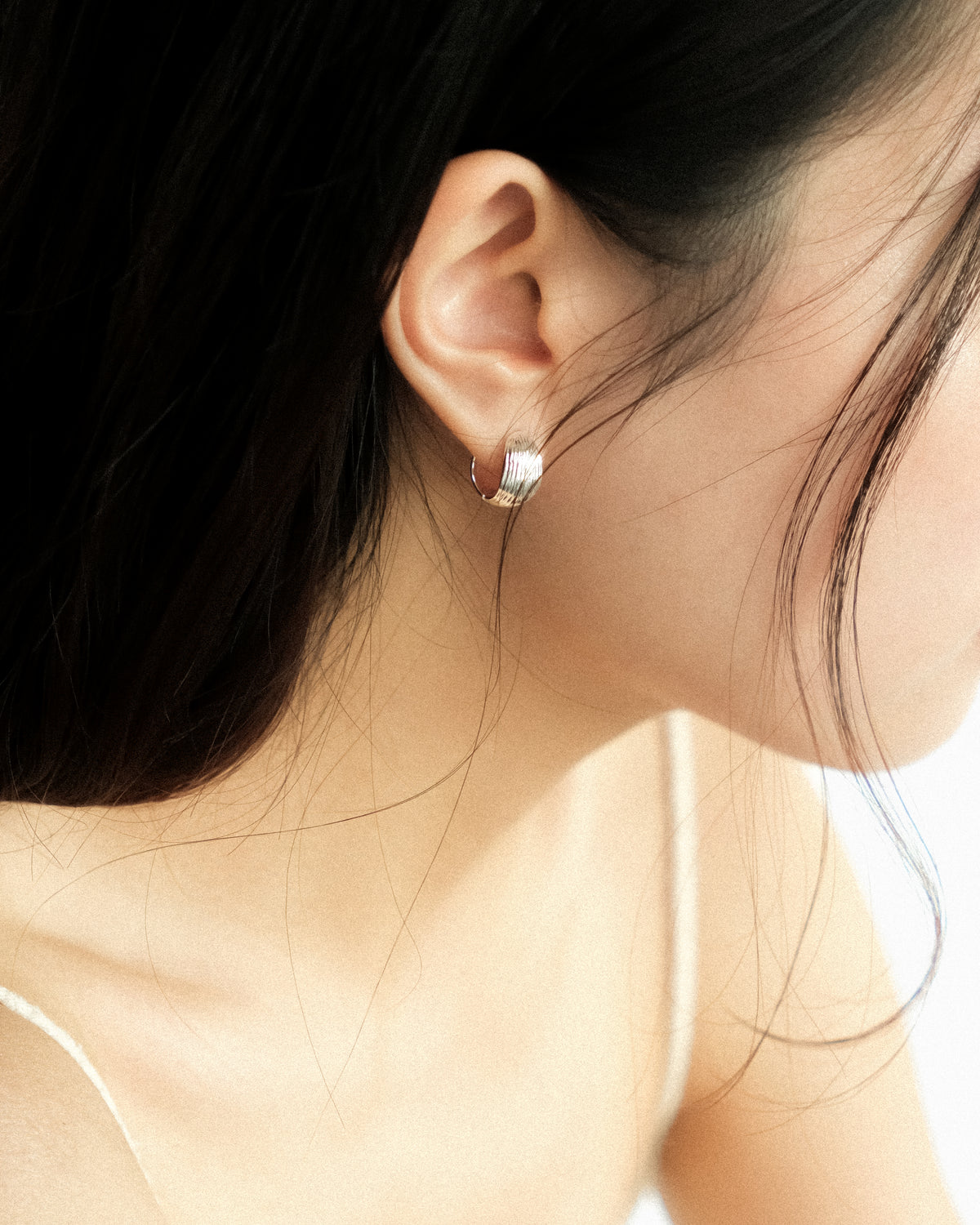 A close-up side profile of a woman wearing a small silver reversible hoop earring, shown against her ear and dark hair.