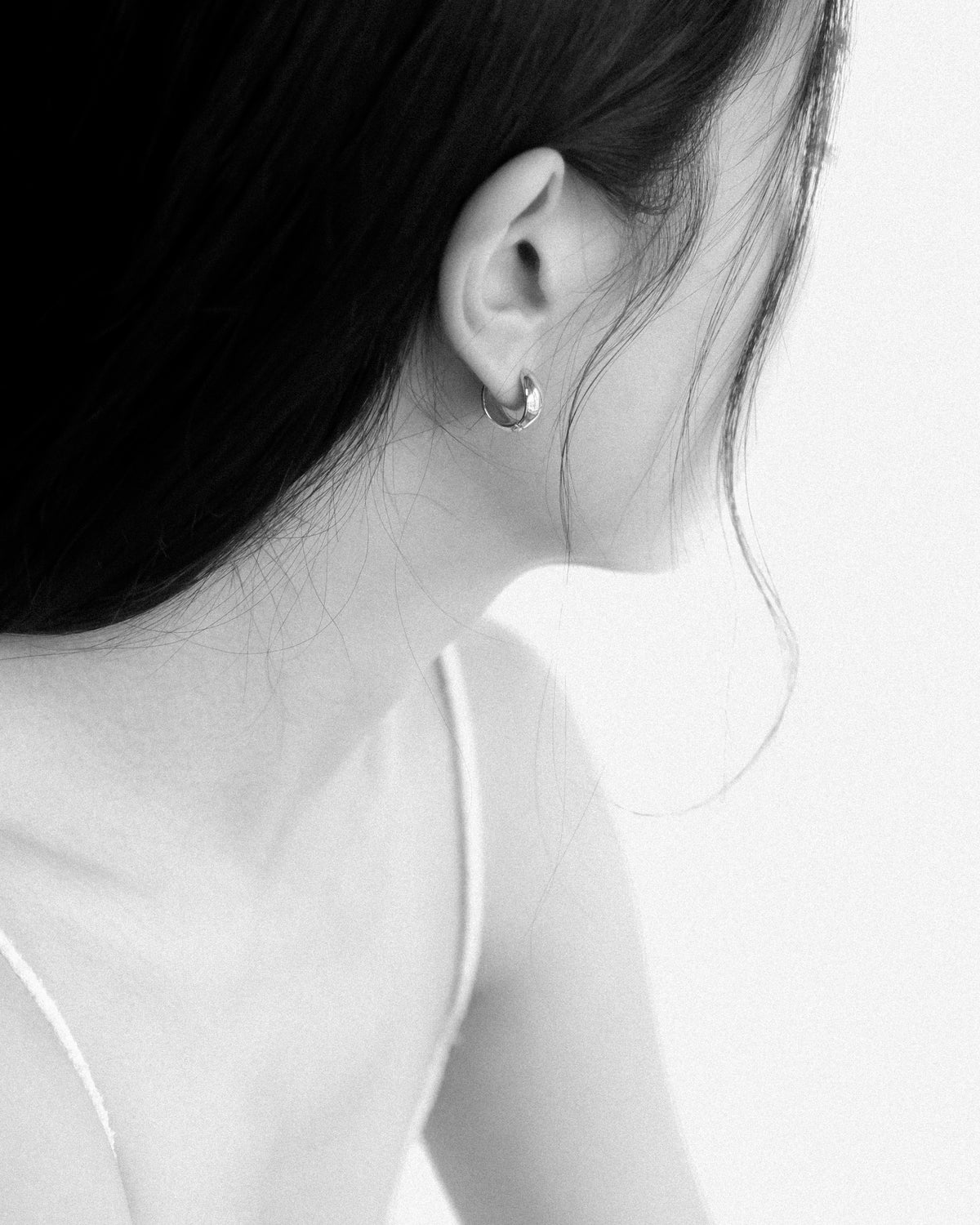 A close-up black and white photograph of a woman's ear wearing a small reversible hoop earring, with her hair and shoulder visible against a plain light background.
