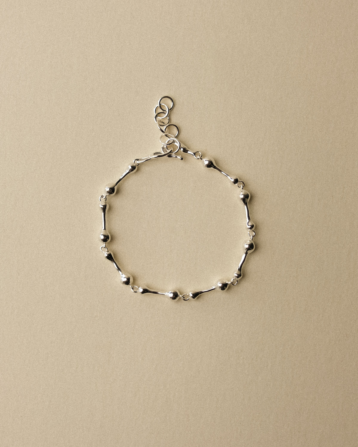 A delicate silver droplet link chain bracelet with alternating polished finishes, completed with a fish hook clasp, displayed against a textured warm wabi-sabi inspired background.