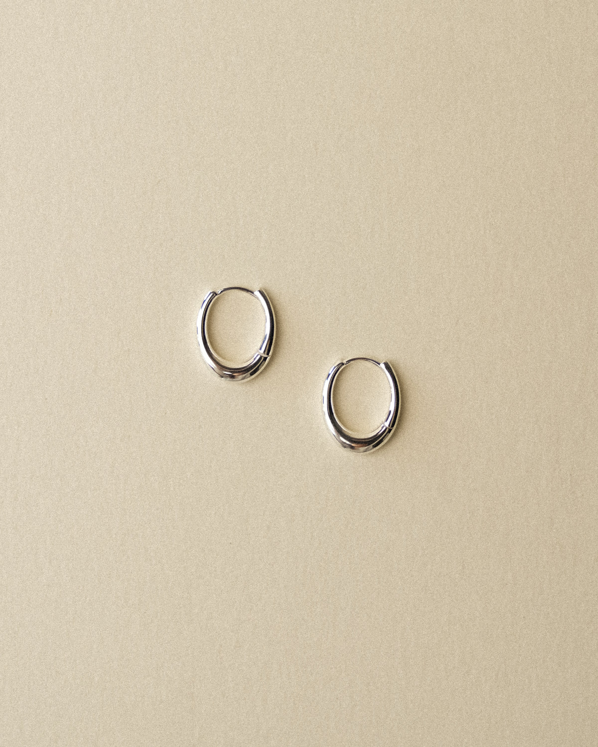 A pair of polished silver pebble shaped hoop earrings displayed on a textured warm wabi sabi background.