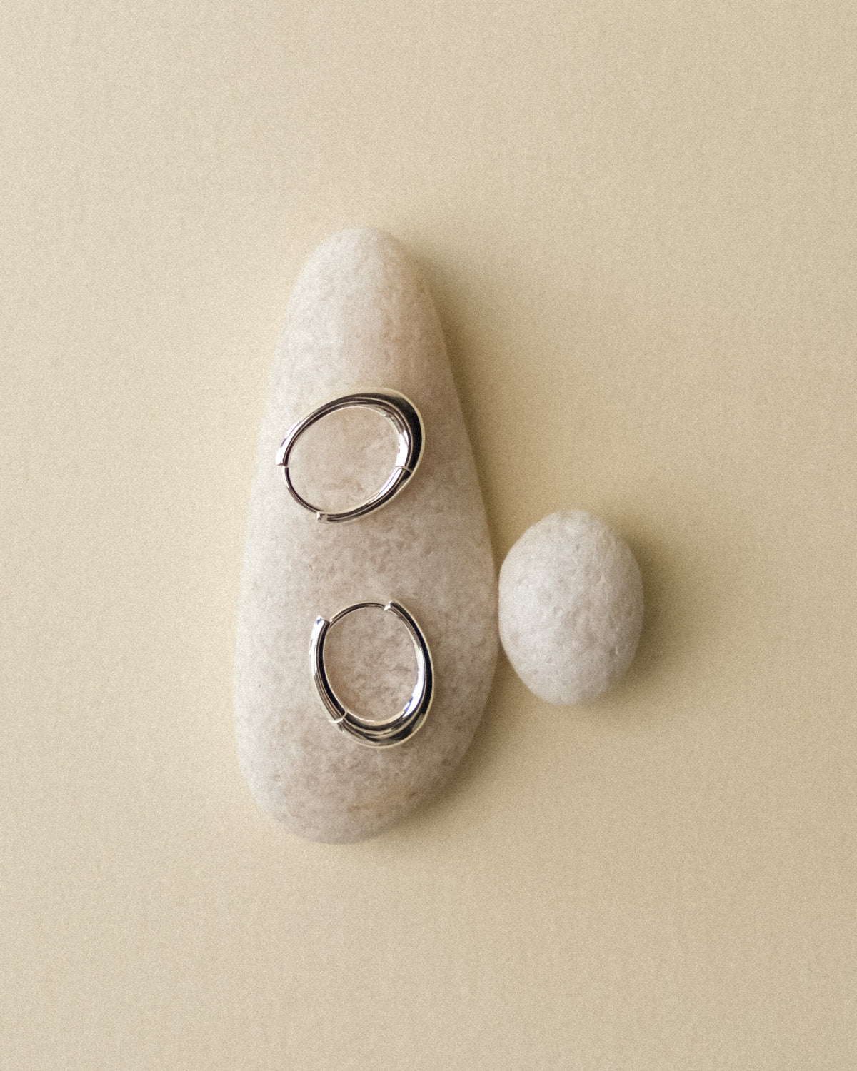 Two silver pebble shaped hoop earrings resting atop white pebbles, echoing their organic form against a warm wabi sabi backdrop.
