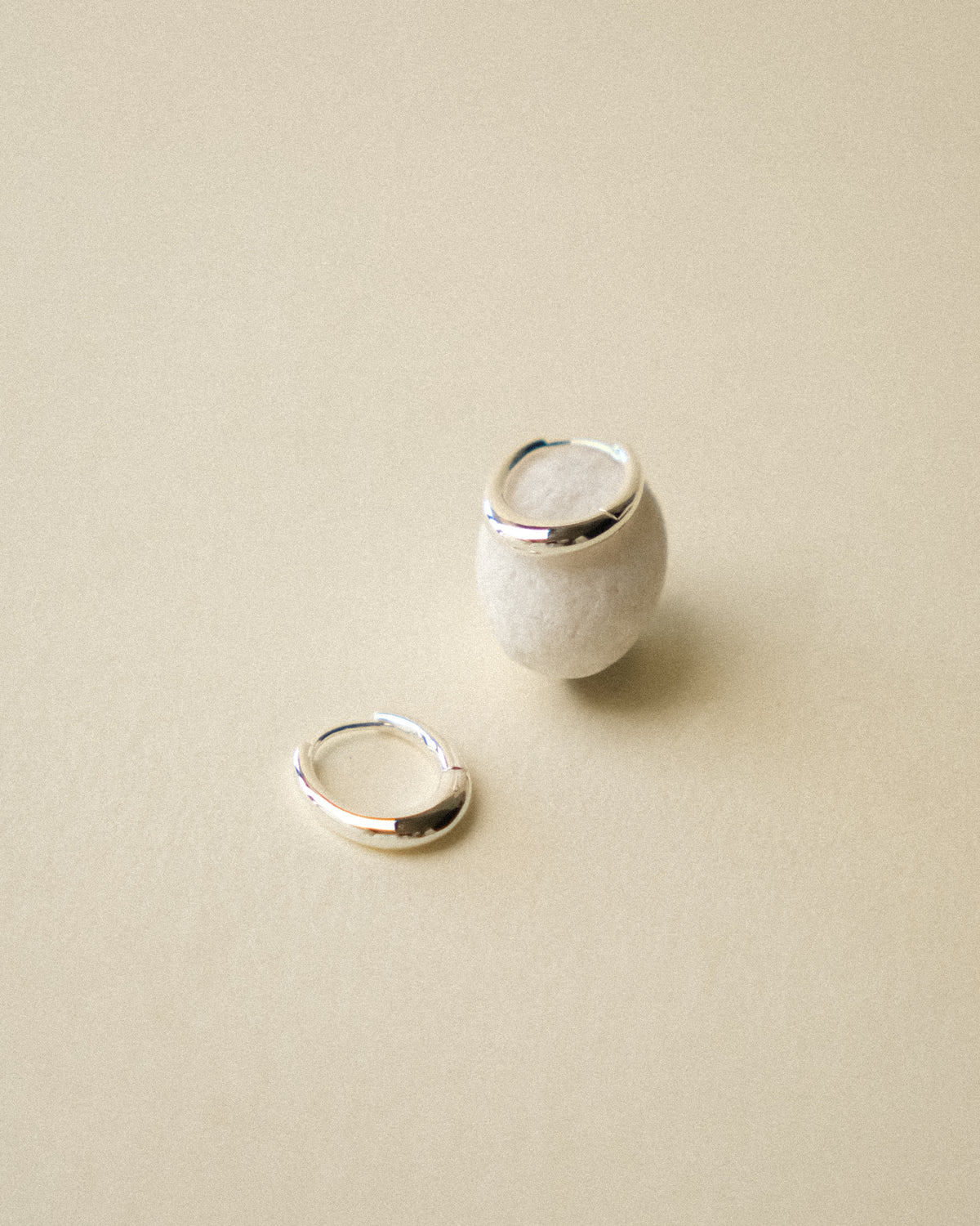 A pair of silver pebble shaped hoop earrings displayed on a warm wabi sabi background, one lying upright on a white pebble stone and one lying flat on the backdrop.