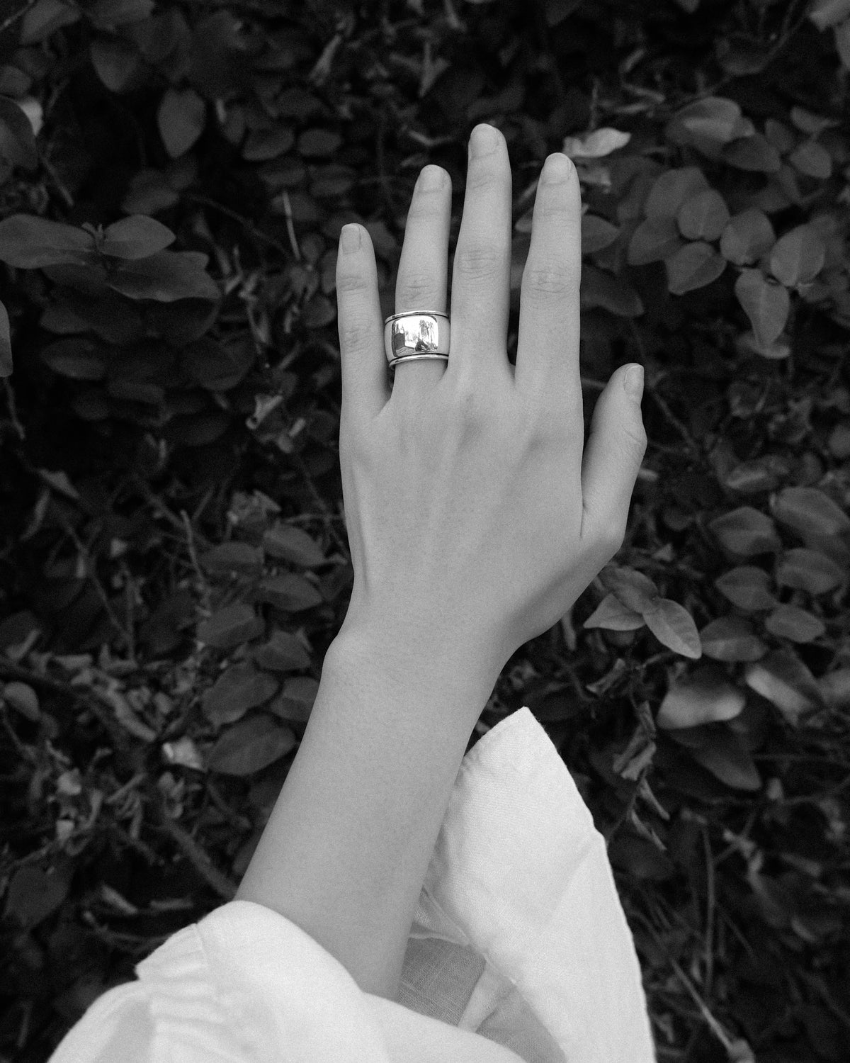A woman wearing a bold sculptural cigar band ring on her wedding finger, photographed against a natural green vine background in monochrome.