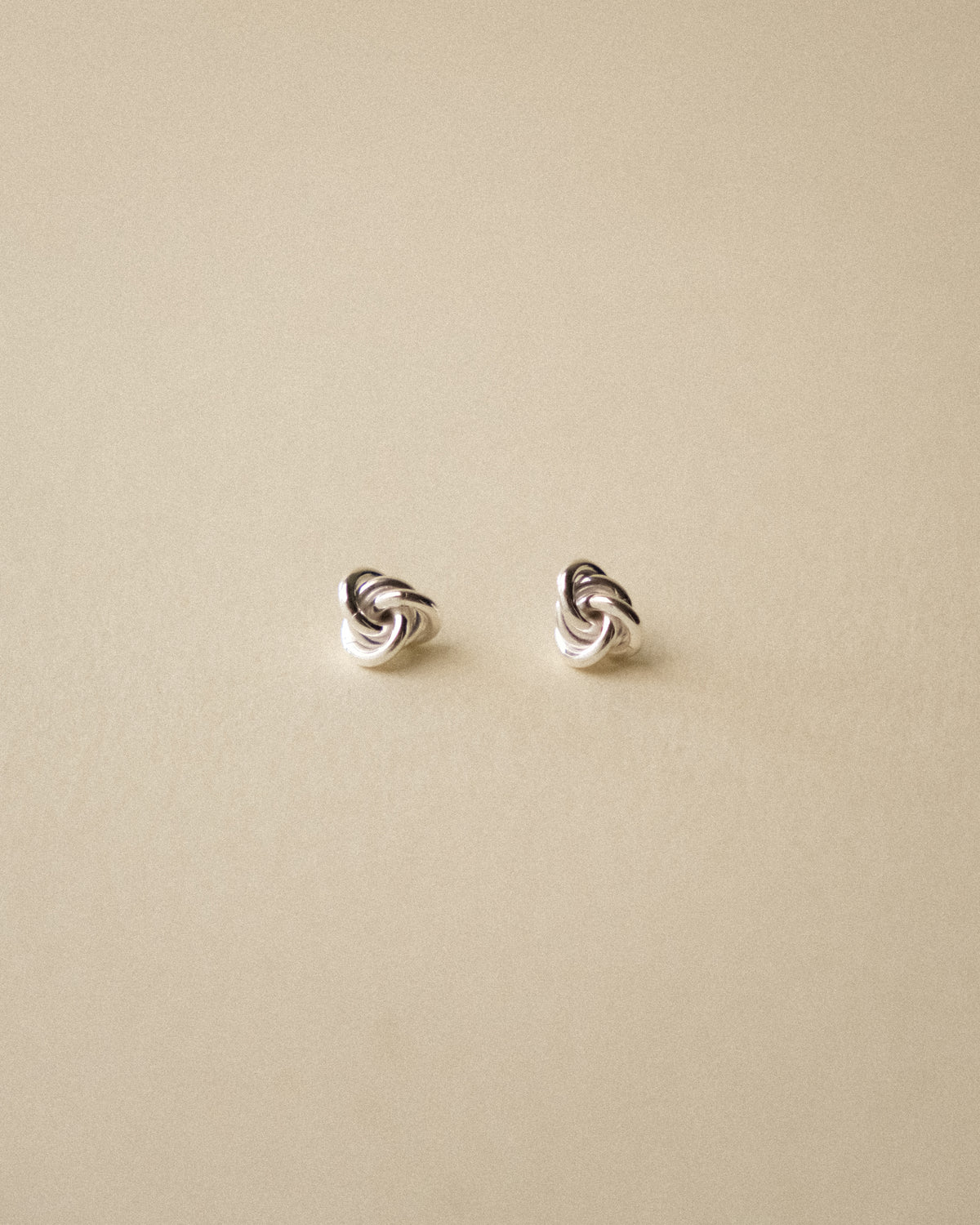 A pair of small silver knot stud earrings on a plain warm wabi-sabi background.