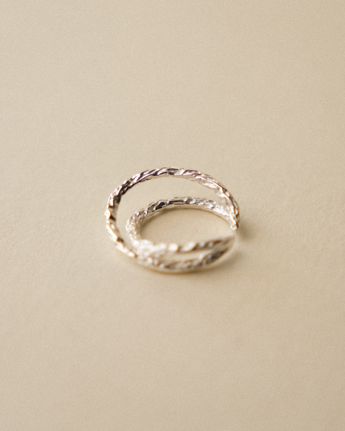 A hammered crossed ring laid flat, captured from the side to highlight its sculptural intersection and handcrafted texture against a warm, minimal wabi-sabi background.