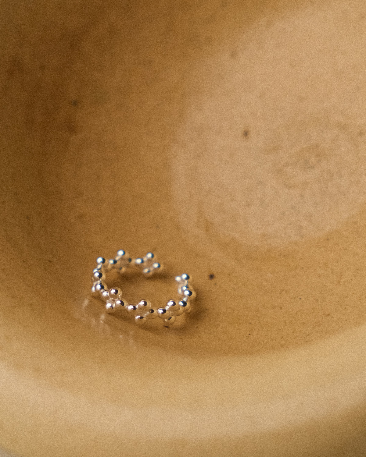 delicate silver floral-shaped ring resting gracefully inside a textured yellow-beige wabi-sabi ceramic bowl. The organic curves of the ring echo the quiet beauty of nature, complemented by the handcrafted imperfections and earthy warmth of the ceramic surface. A minimalist still-life composition with soft natural light, highlighting the subtle texture of silver and the serene elegance of wabi-sabi aesthetics.