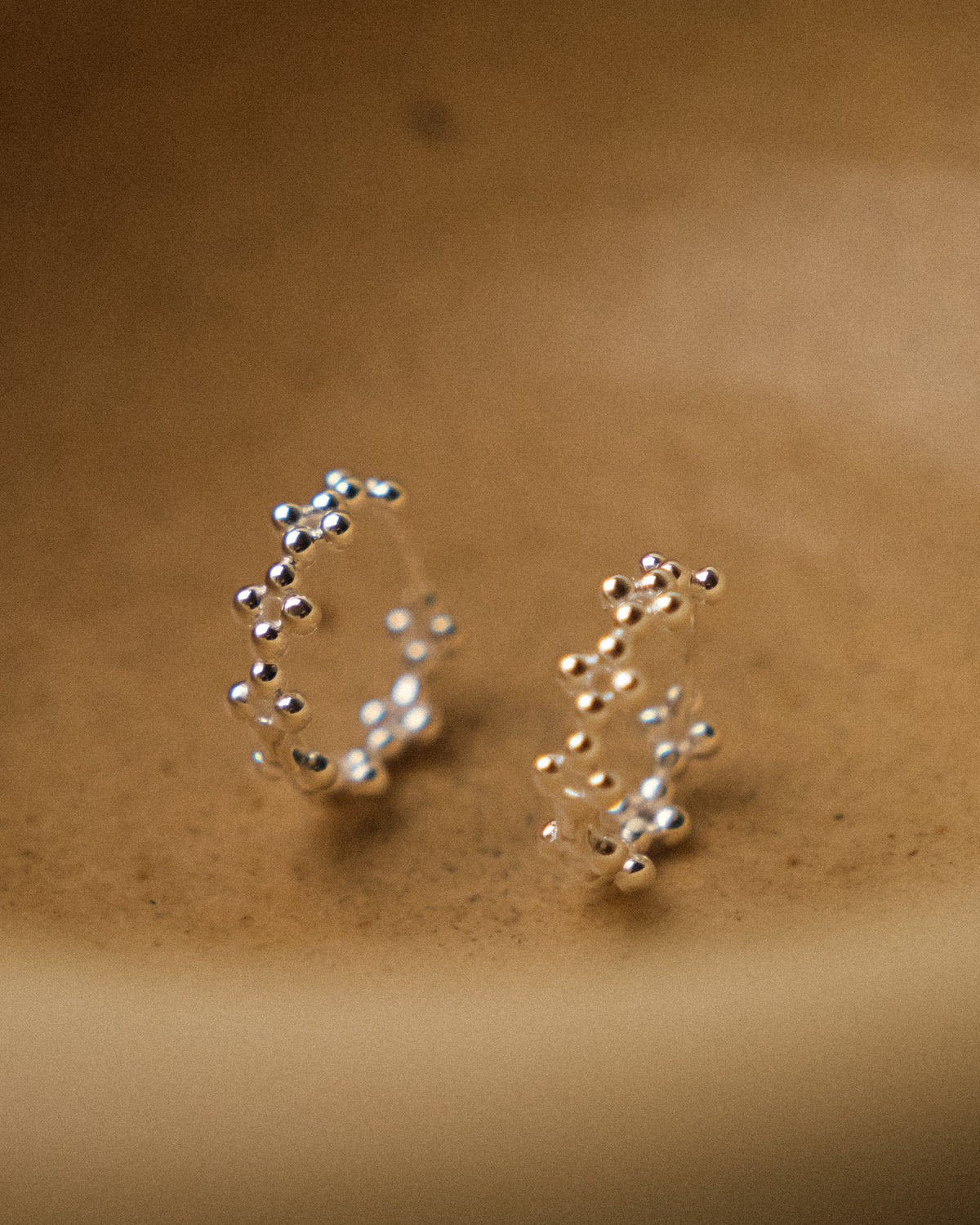 A delicate pair of silver floral-shaped hoop earrings with intricate silver bead details, standing gracefully inside a textured yellow-beige wabi-sabi ceramic bowl. The organic curves of the earrings echo the quiet beauty of nature, complemented by the handcrafted imperfections and earthy warmth of the ceramic surface. A minimalist still-life composition with soft natural lighting, highlighting the subtle textures of silver, the sculptural floral form, and the serene elegance of wabi-sabi aesthetics.