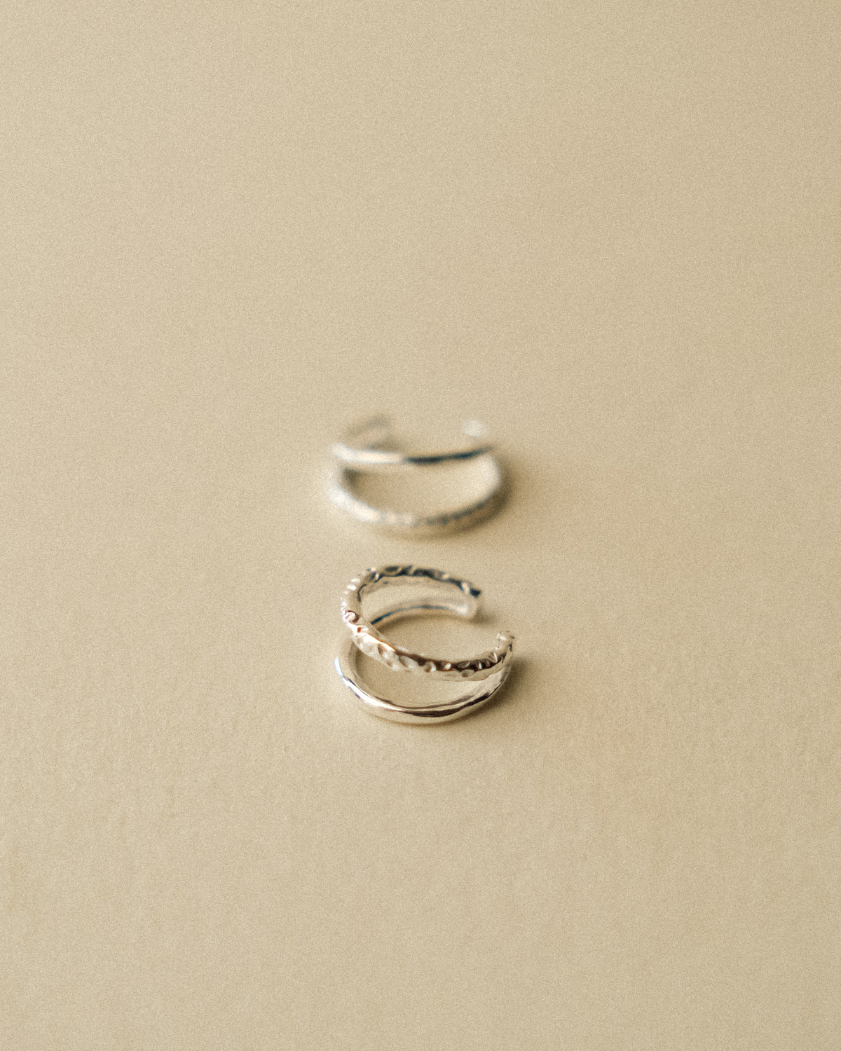 A pair of silver ear cuffs embracing the ear with a double-layered design, balancing the raw texture of hand-hammered silver with the refined sheen of a polished finish, captured from the front with focus on the first ear cuff against a warm wabi-sabi inspired background.