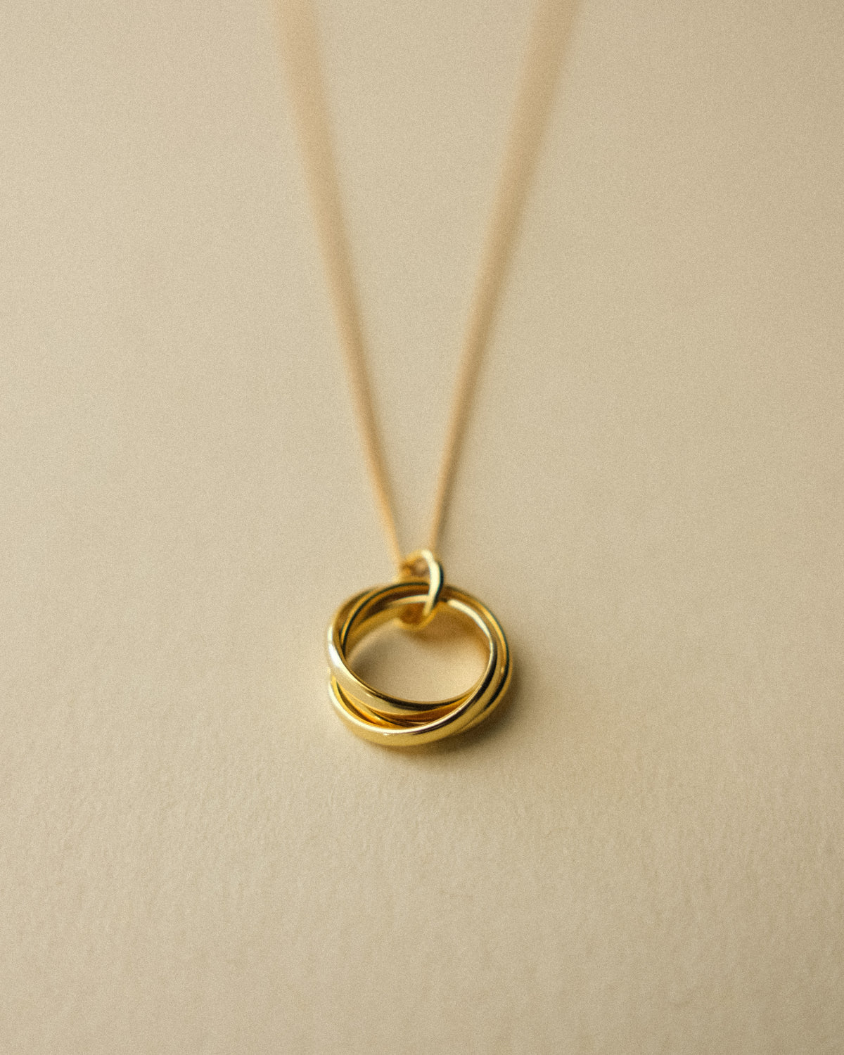 A gold Trio Interlink pendant necklace captured from the front, set against a warm wabi-sabi backdrop that highlights its sculptural and timeless form.