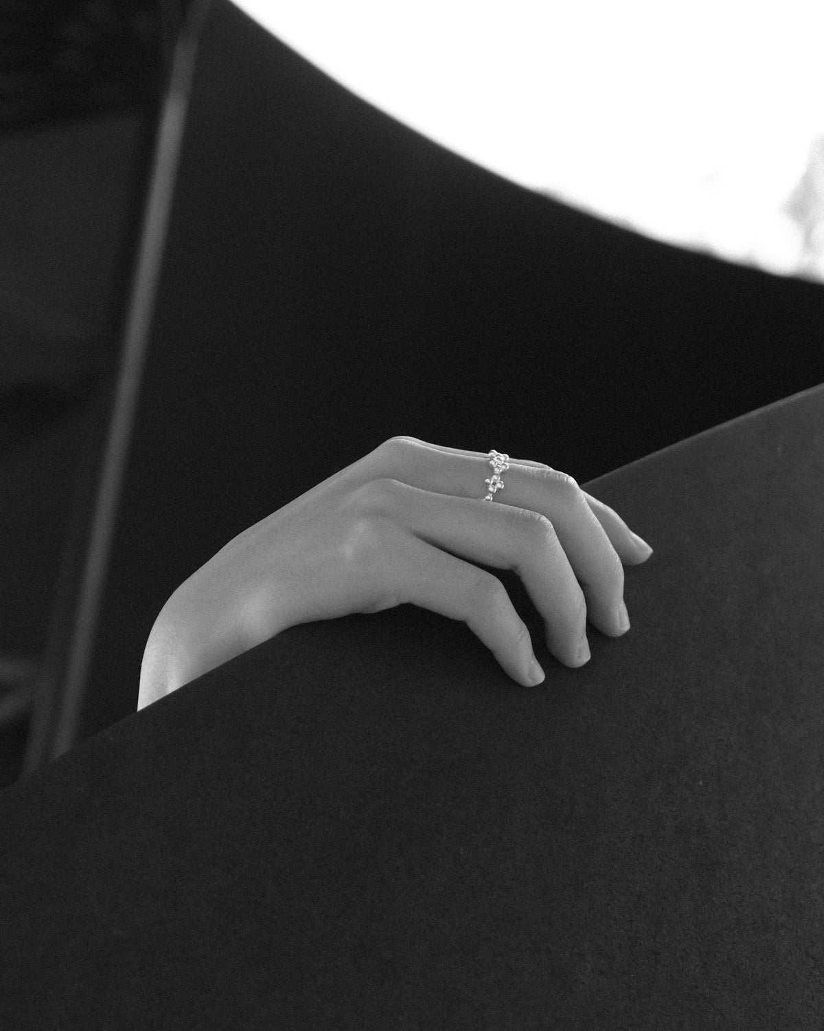 A close-up black and white photograph of a person’s hand resting gracefully along a spiral staircase, adorned with a delicate silver floral-shaped ring. The flowing curves of the staircase echo the organic form of the jewellery, creating a harmonious dialogue between architecture and craftsmanship. Soft monochrome tones highlight the intricate details of the silver, the texture of the hand, and the sculptural elegance of the surrounding space.