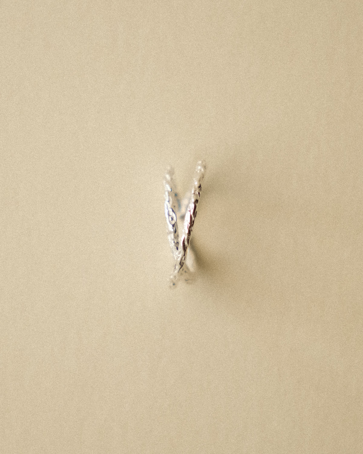 A front-facing shot of the hammered crossed ring, highlighting its sculptural intersection and textured details against a warm, minimal wabi-sabi background.