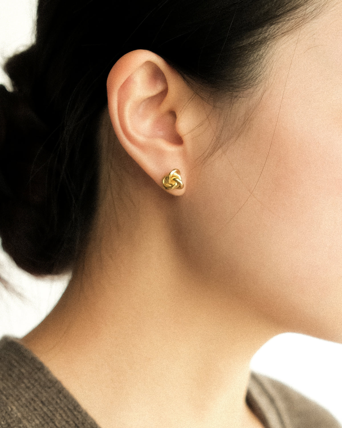 A close-up side profile of a woman's ear wearing a small gold knot stud earring.