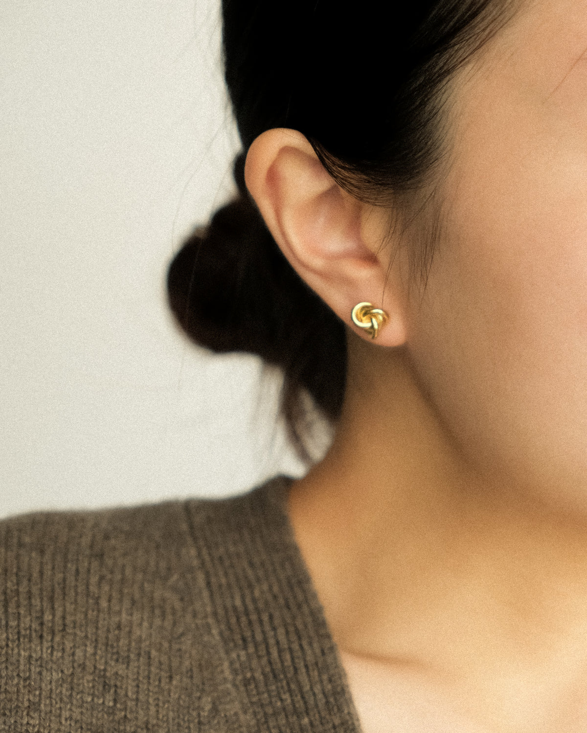 A close-up of a person's ear wearing a small gold twisted knot stud earring, with dark hair tied back and a knit brown top against a plain light background.