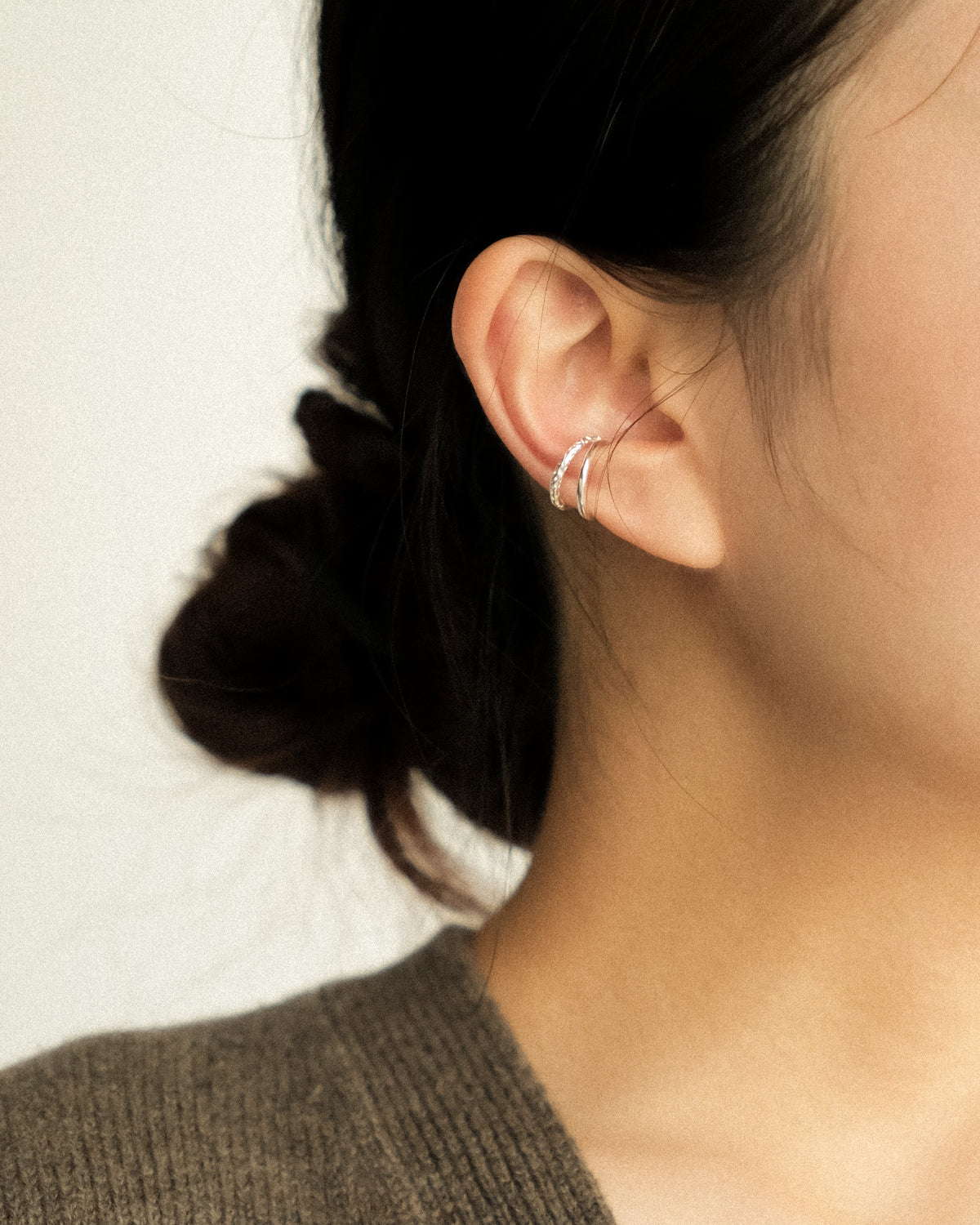 A woman wearing a silver ear cuff with a double-layered design, balancing the raw texture of hand-hammered silver with the refined sheen of a polished finish, captured in a side profile against a clean white background.