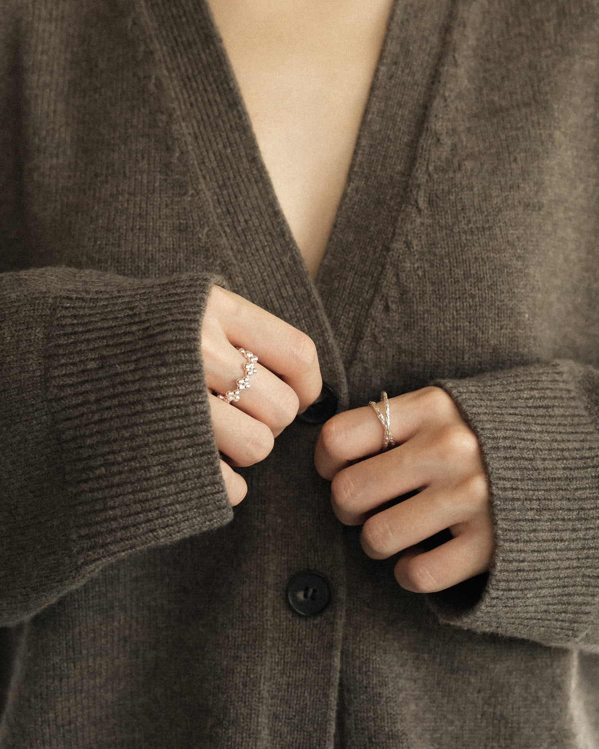 A woman wearing a soft V-neck wool knit outer in brown, gently opening the buttons with both hands. Her hands are adorned with a delicate silver floral-shaped ring paired with a crossover hammered line ring, creating a sculptural and harmonious jewellery pairing. A quiet, minimalist fashion scene highlighting the organic textures of wool, handcrafted silver details, and the subtle elegance of everyday styling.
