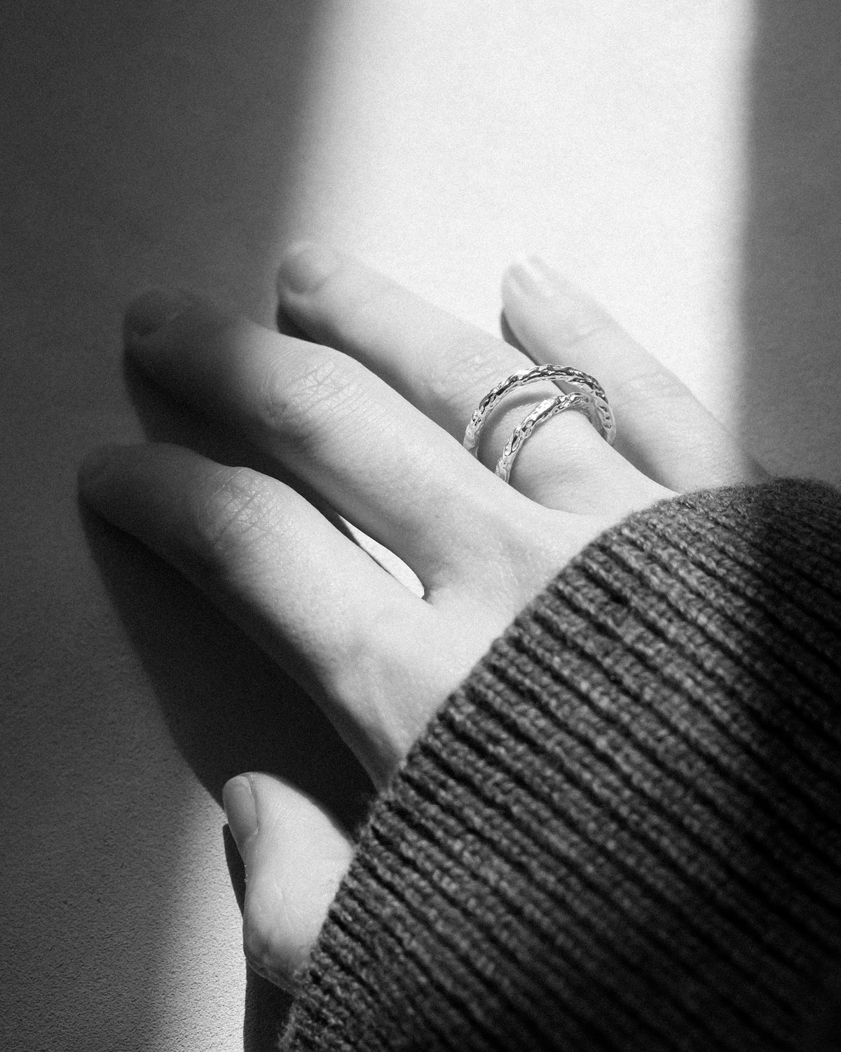 A close-up black and white photograph of a person's hand resting on a surface, wearing a hammered crossed rings. The image is shot in high contrast with dramatic shadows and highlights.