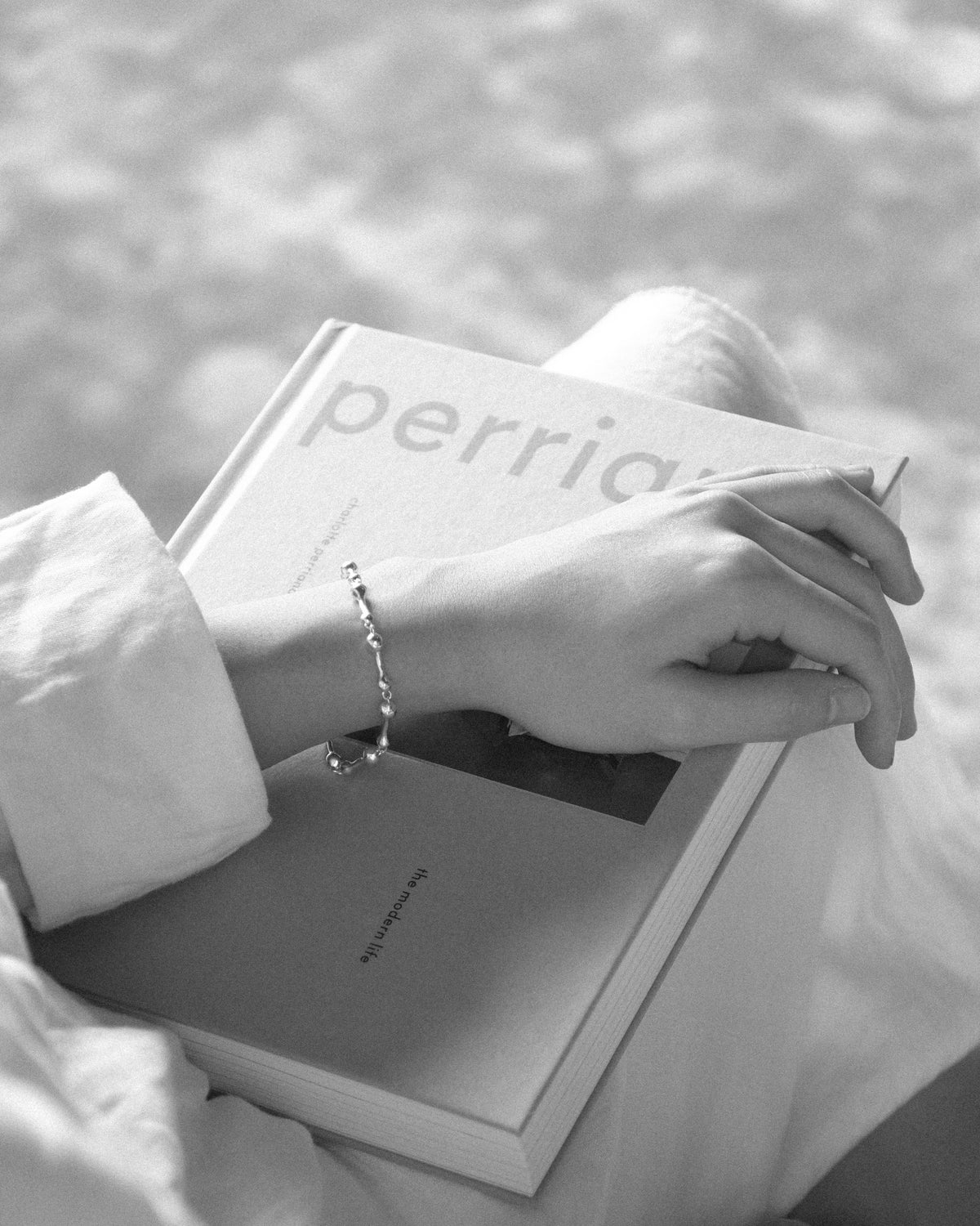 A close-up black and white photograph of a person's hand resting on a book, wearing a delicate silver droplet link chain bracelet. The person is dressed in a white long-sleeve top, and the background is a soft-focus setting.