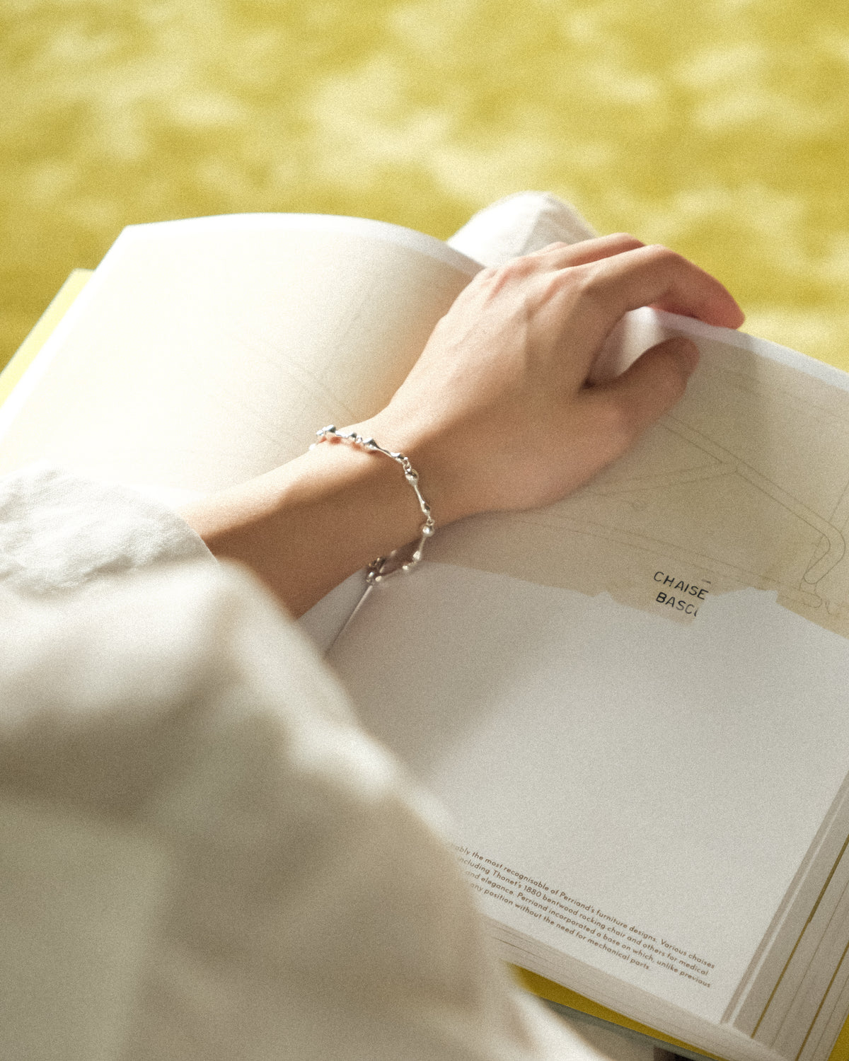 A close-up lifestyle shot of a person's hand resting on an open book, wearing a delicate silver droplet link chain bracelet.