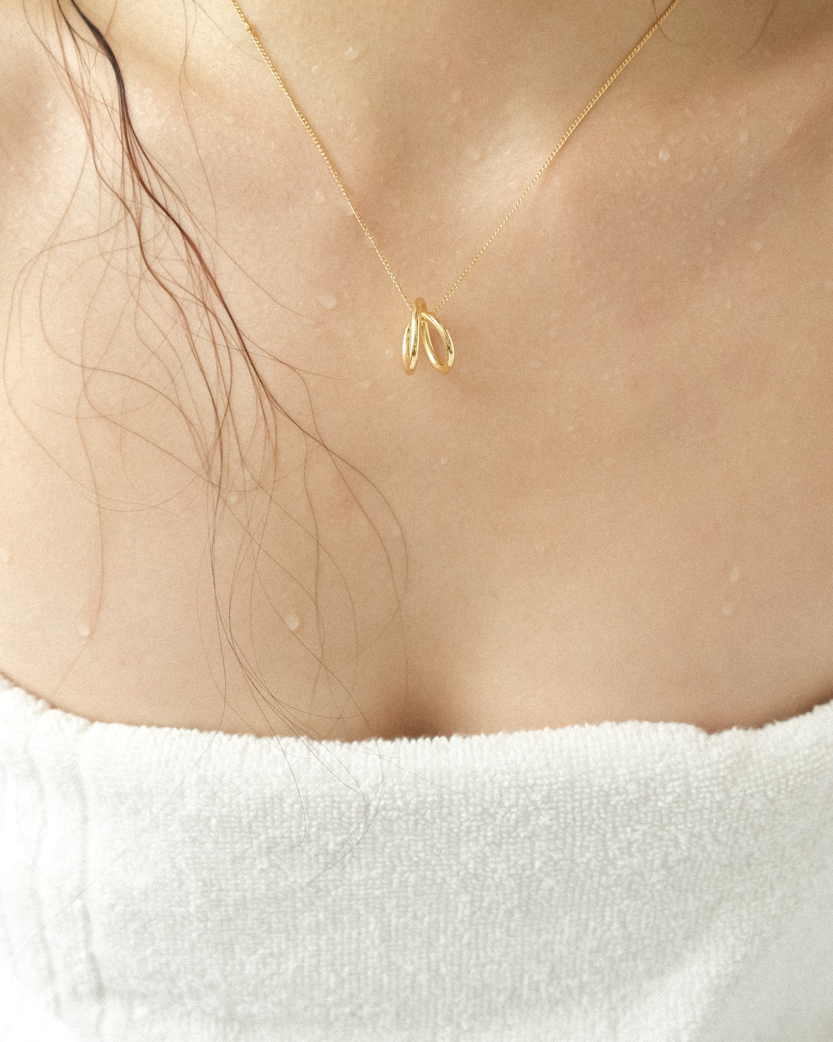 A gold line sculpture pendant necklace worn by a person wrapped in a bath towel.
