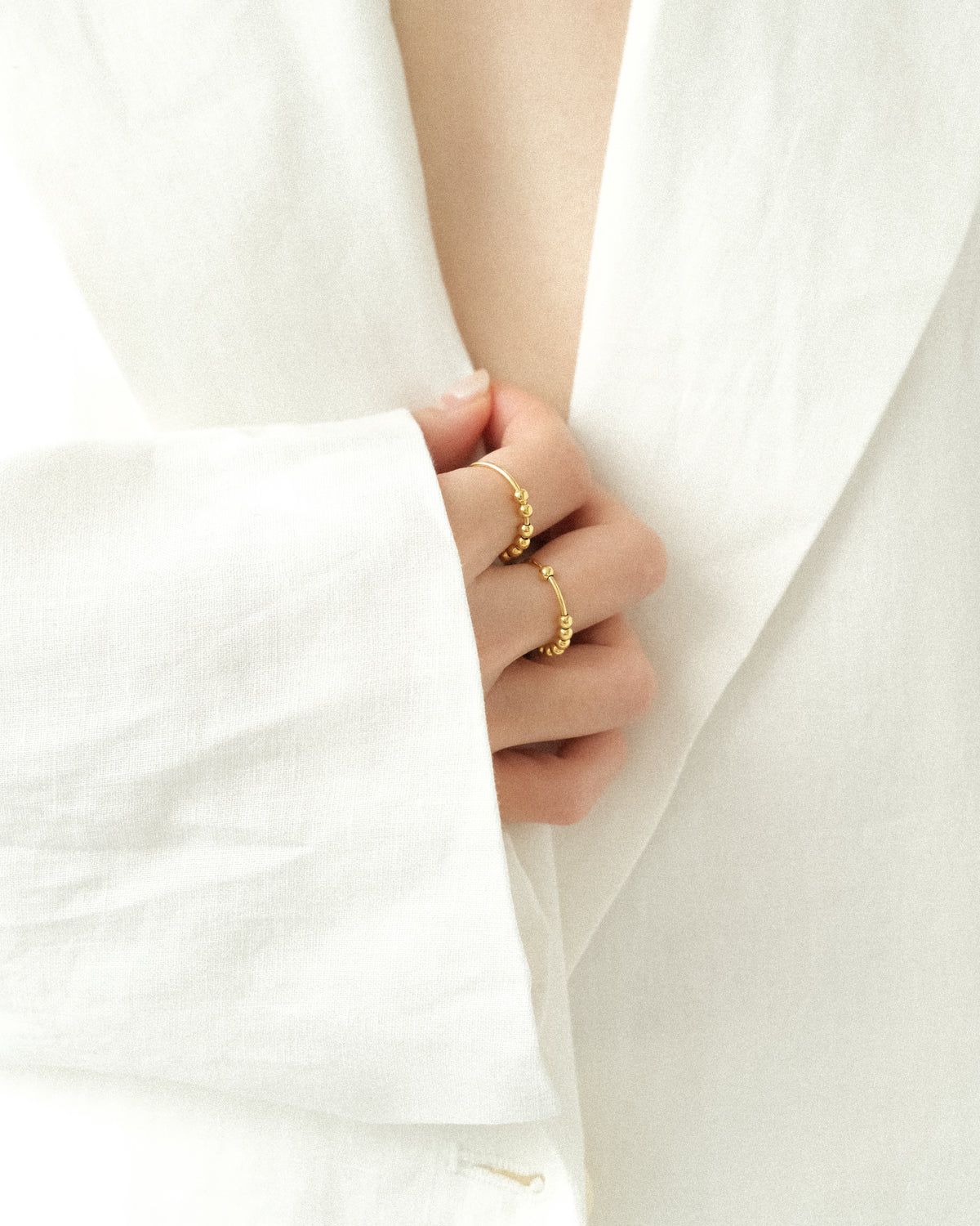 A person in a white linen coat, captured in a close-up focusing on the hand, wearing gold vermeil rings on the index and middle fingers, refined and understated with a soft natural light.