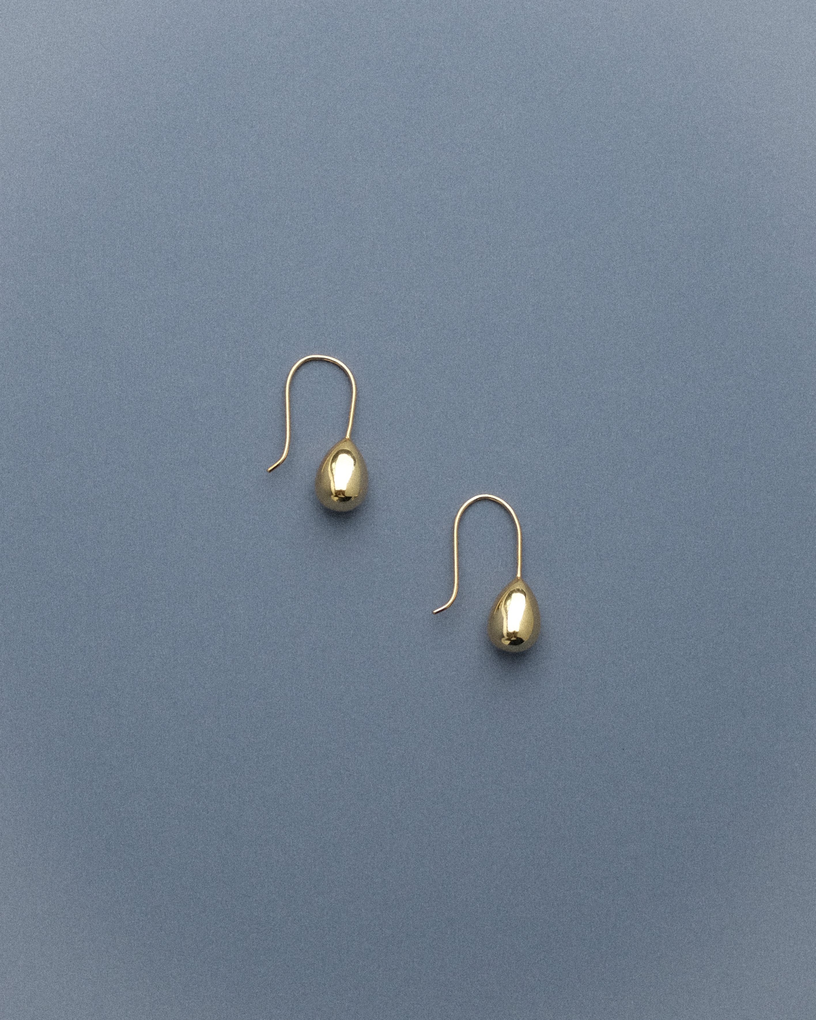 A pair of gold egg drop dangle earrings captured from the side, resting against a soft grey-blue background, their sculptural curves catching the light gently.