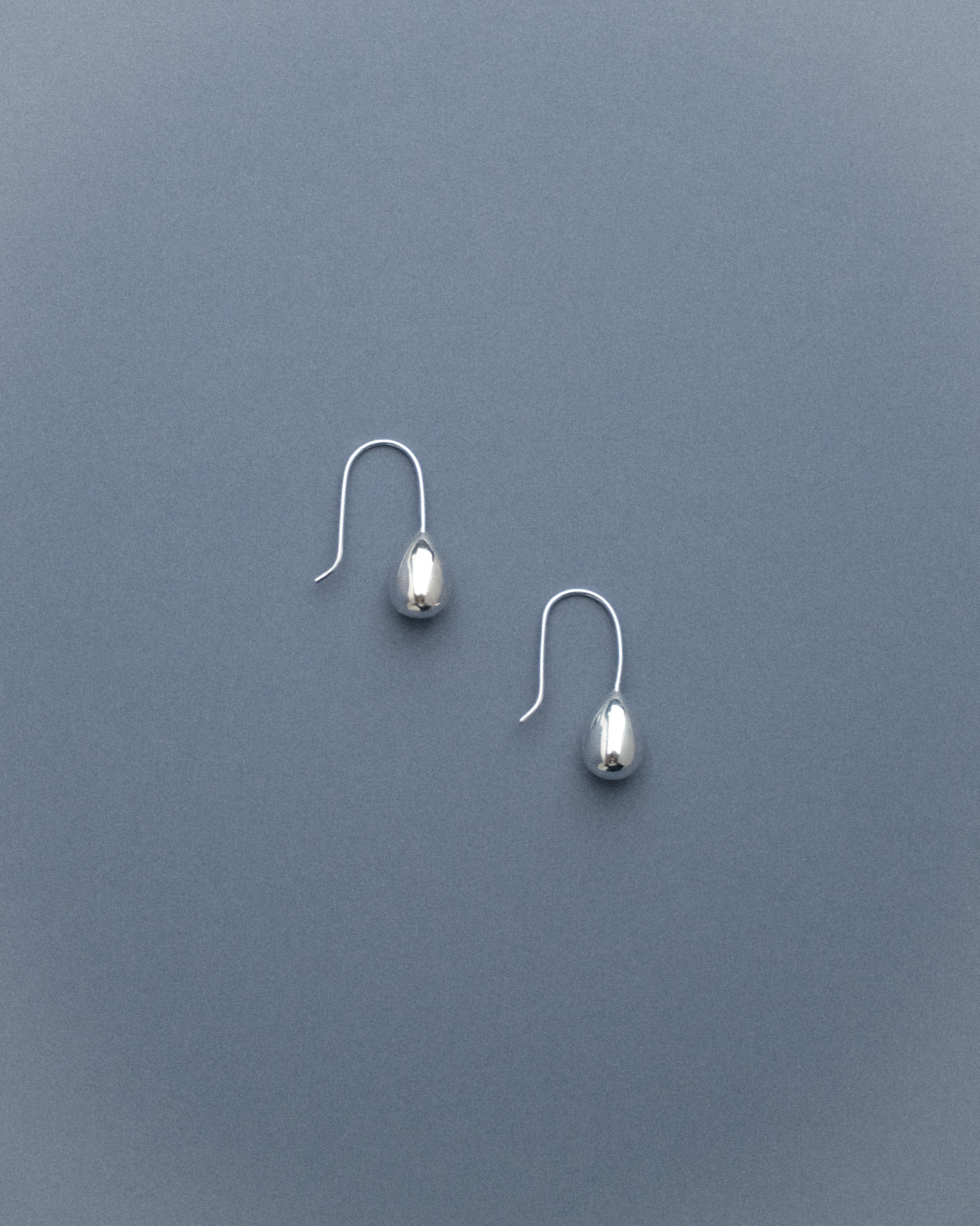 A pair of silver egg drop dangle earrings captured from the side, resting against a soft grey-blue background, their sculptural curves catching the light gently.