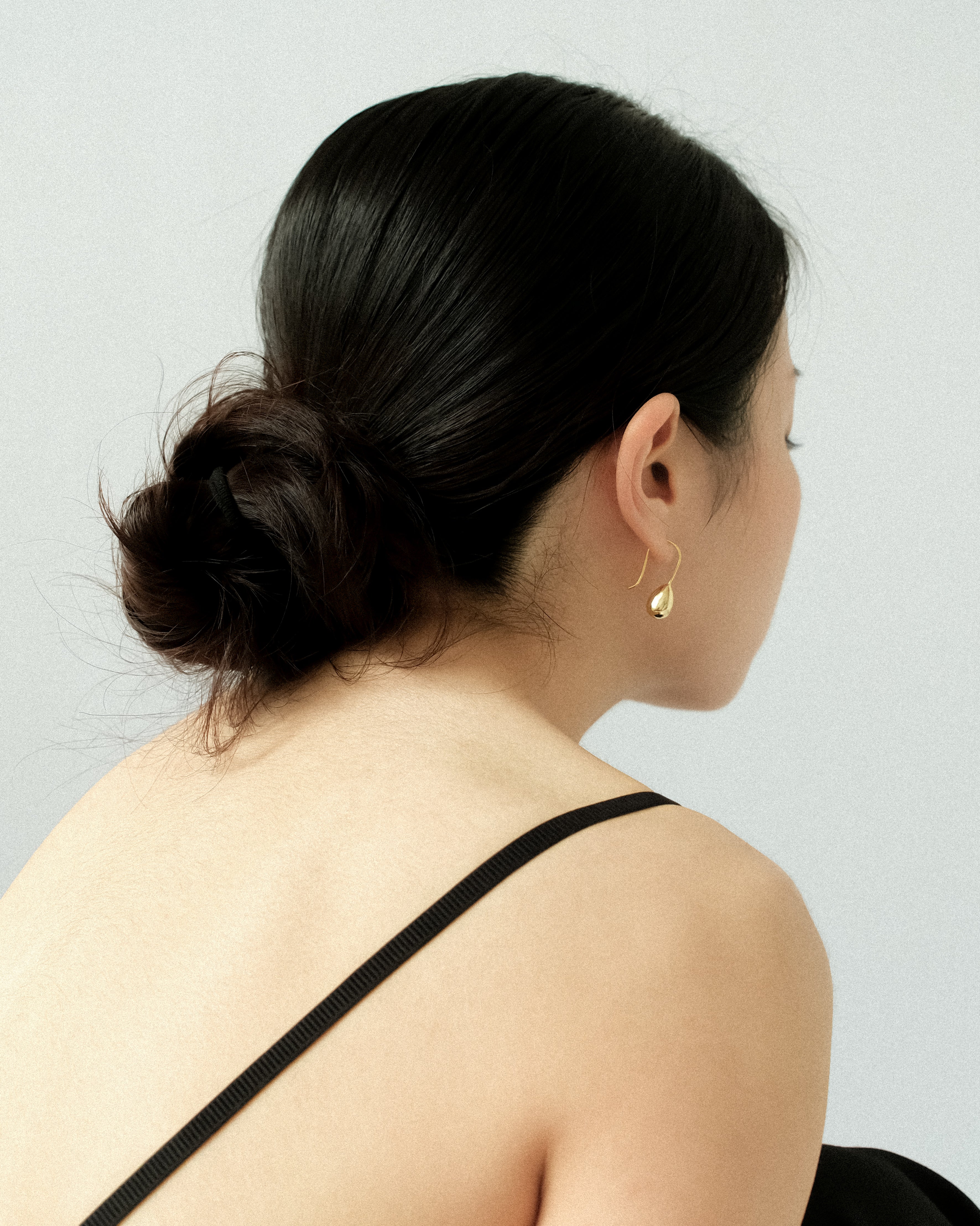 A woman with a sleek hair bun, wearing a black singlet top, adorned with a pair of gold egg drop dangle earrings, standing against a plain, minimalist background.