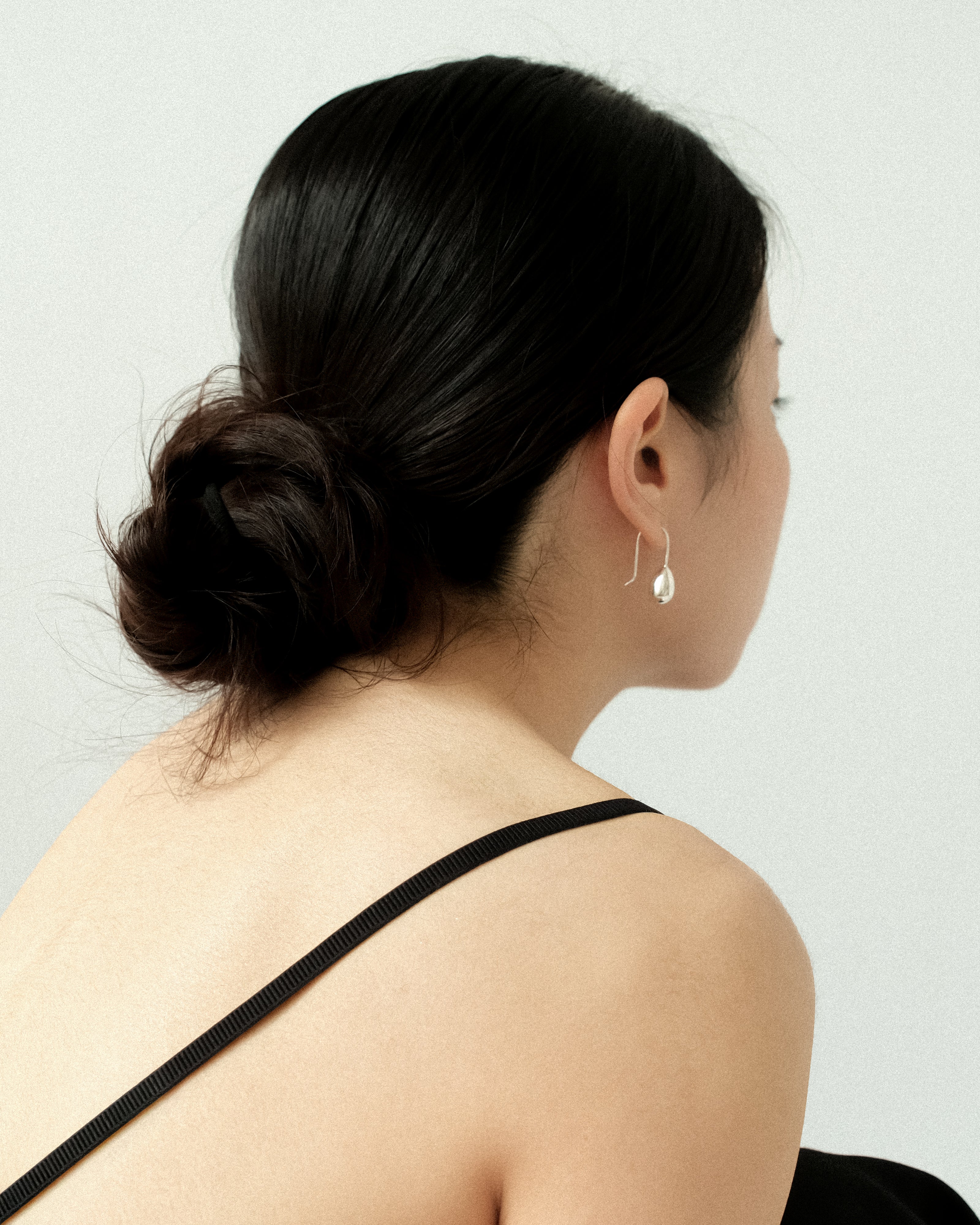 A woman with a sleek hair bun, wearing a black singlet top, adorned with a pair of silver egg drop dangle earrings, standing against a plain, minimalist background.