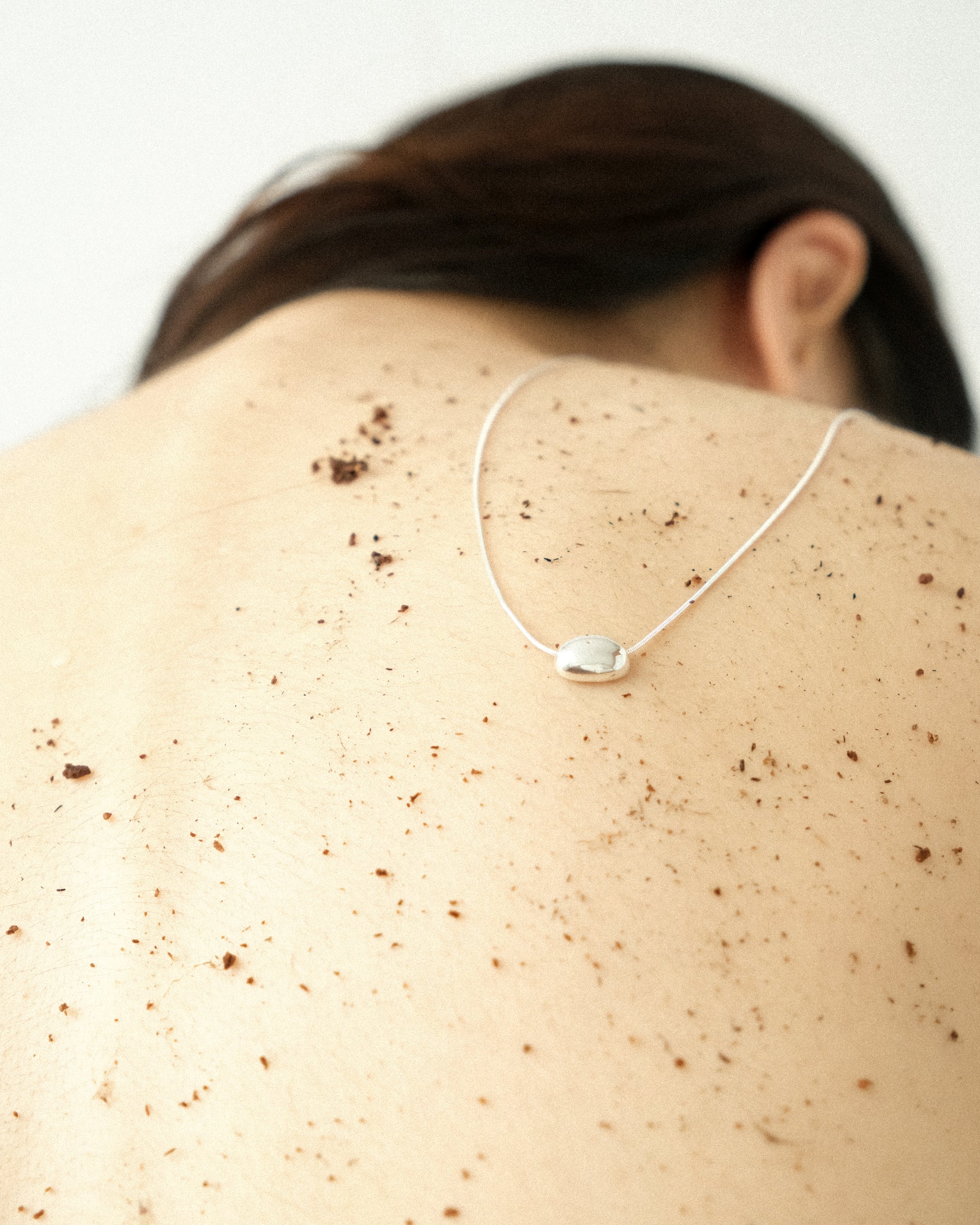 A woman shot from the back, wearing a silver egg-form necklace resting against her skin, dusted lightly with soil, set against a stark white background.