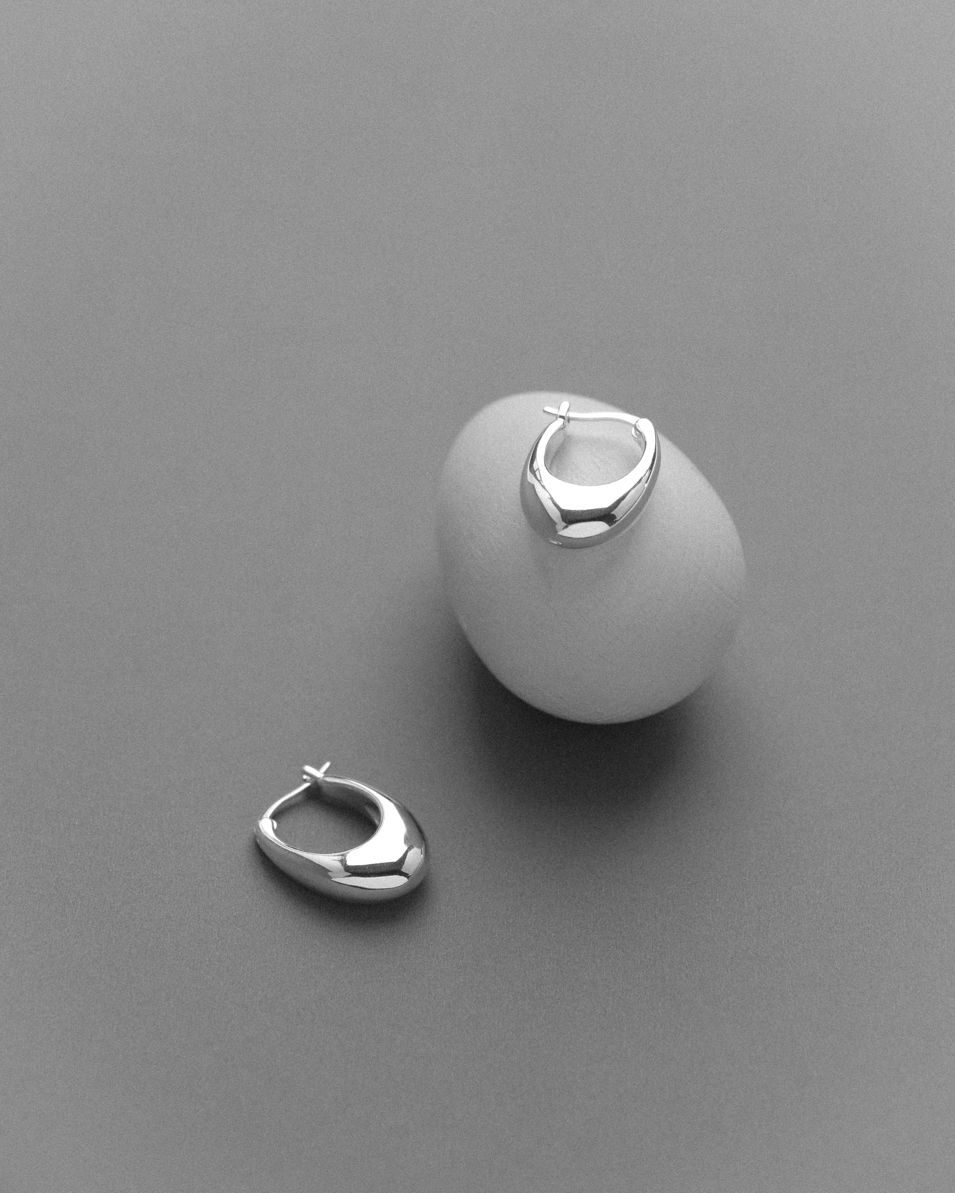 A pair of silver hoop earrings, arranged singly on an egg-like form, photographed against a soft grey surface.