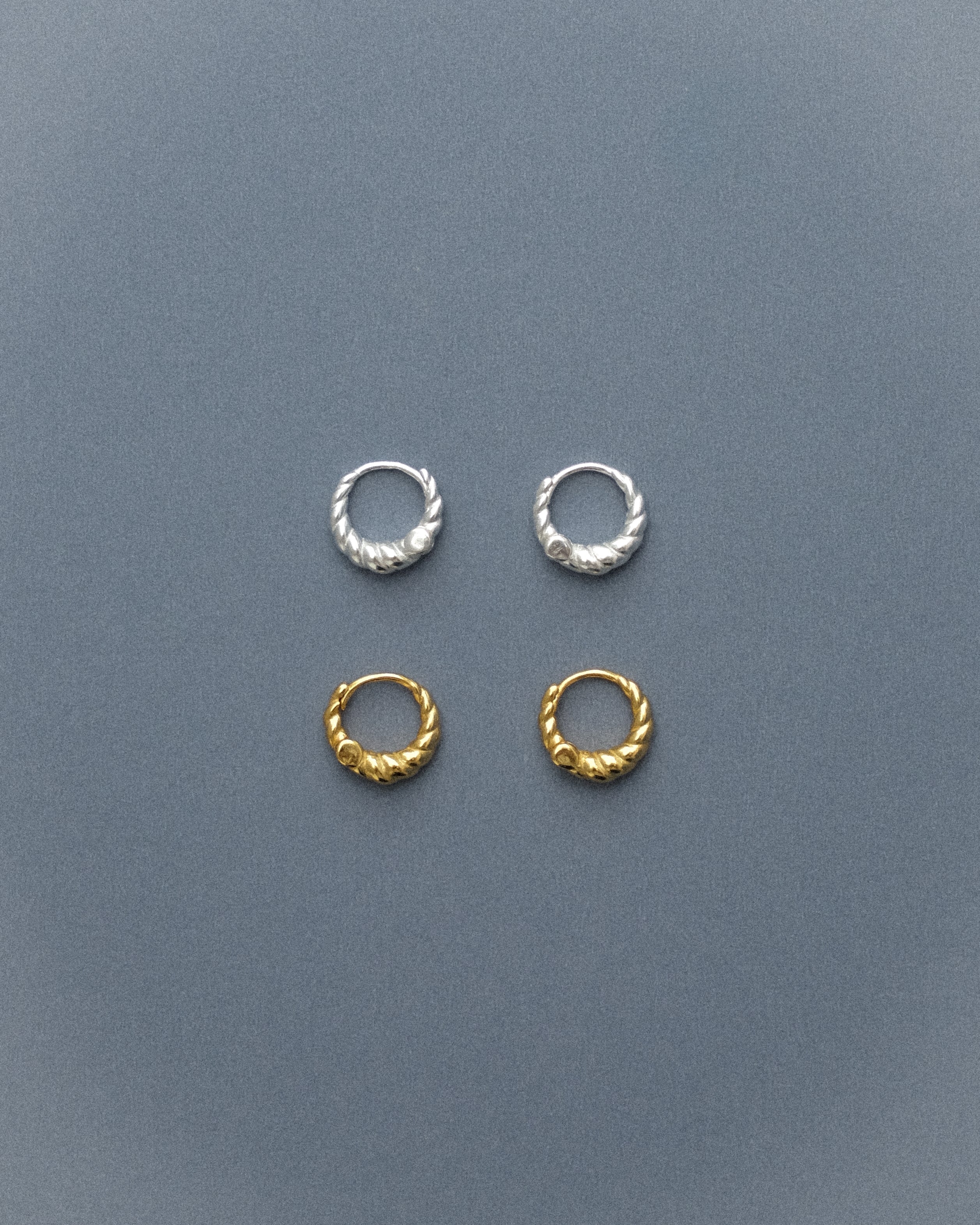 Two pairs of spiral hoop earrings, one in gold and one in silver, arranged in a delicate flatlay, their tiny forms resting on a quiet grey-blue background.