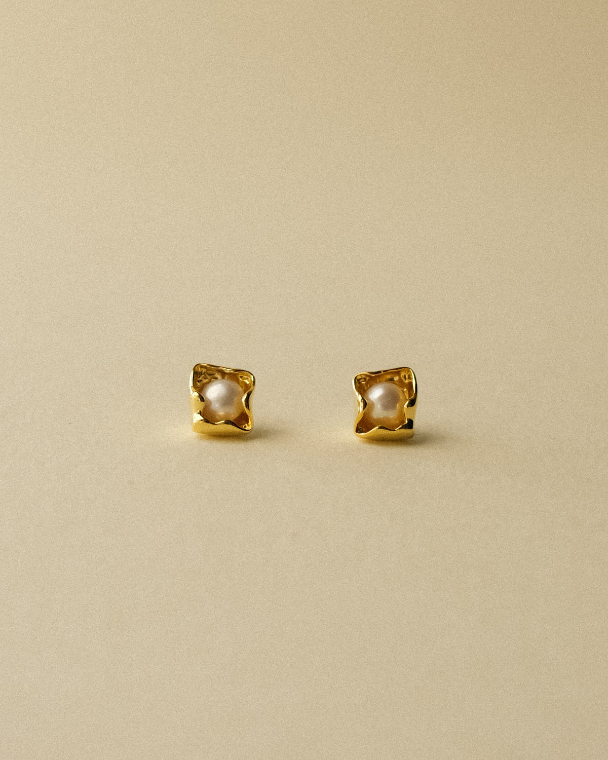 A pair of petal-shaped gold stud earrings with a central white pearl, set against a wabi sabi warm background.