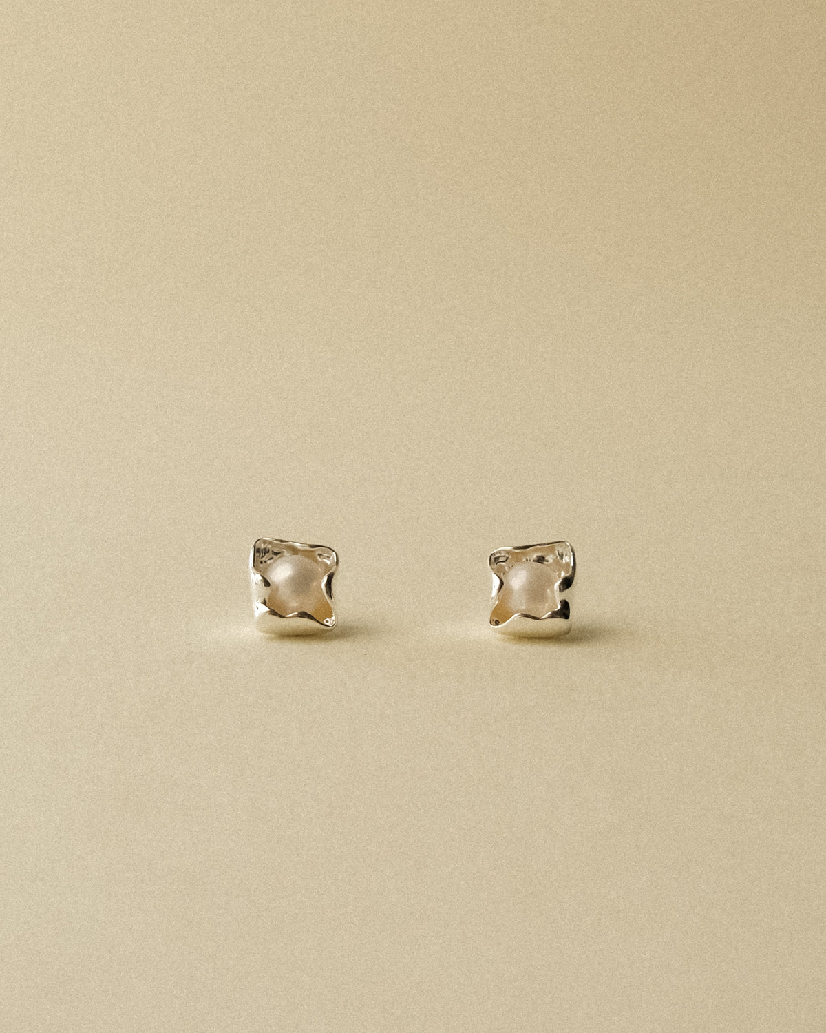A pair of petal-shaped silver stud earrings with a central white pearl, set against a wabi sabi warm background.