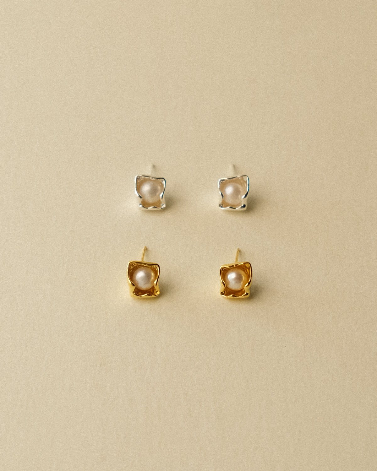 A front-facing shot of a pair of gold and silver petal-shaped earrings, highlighting their geometric form and distinctive white freshwater pearls, set against a warm Wabi-Sabi-inspired background.