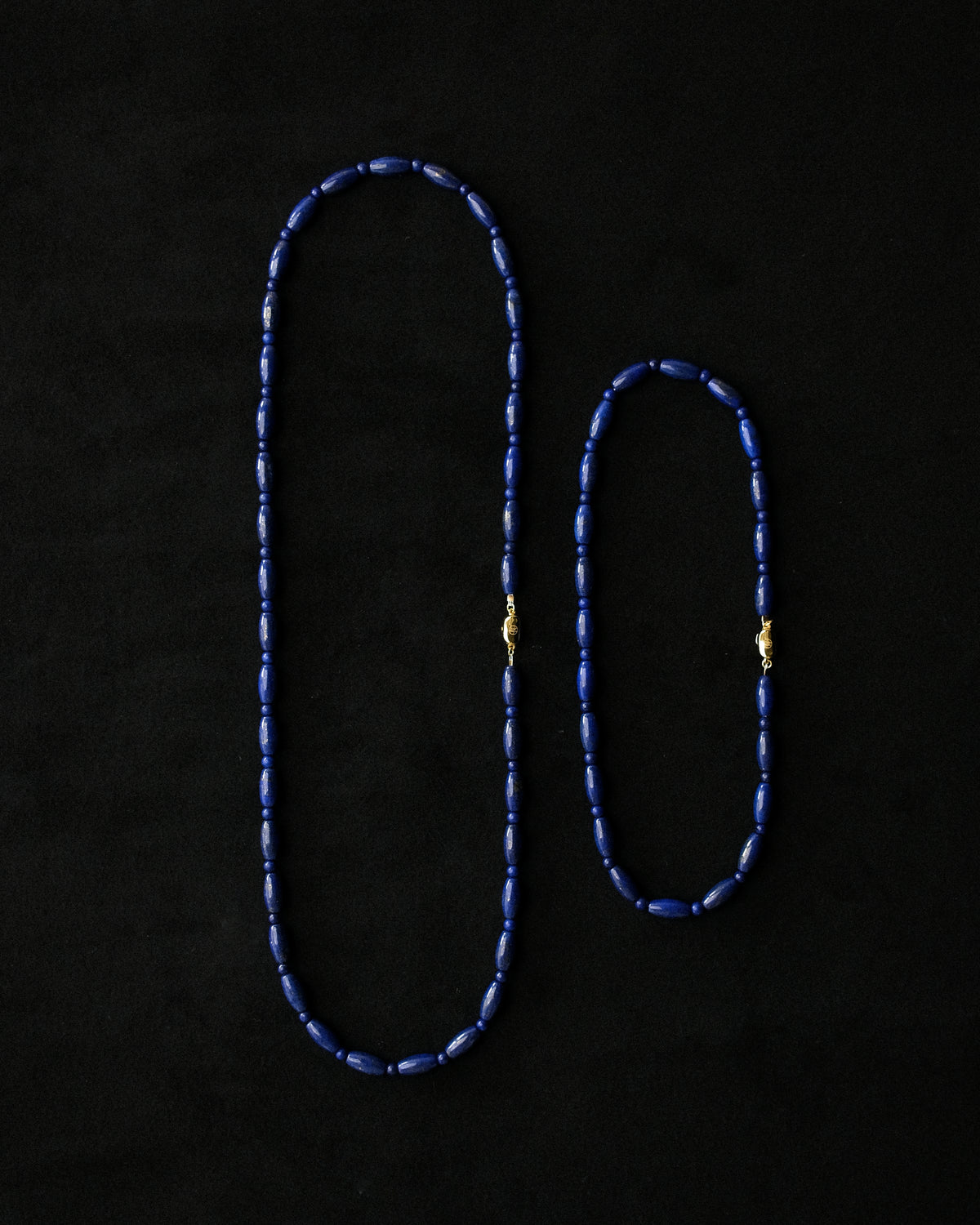 A beaded necklace and choker laid flat against a black background, crafted from small dark blue lapis beads with delicate gold-toned connector details.