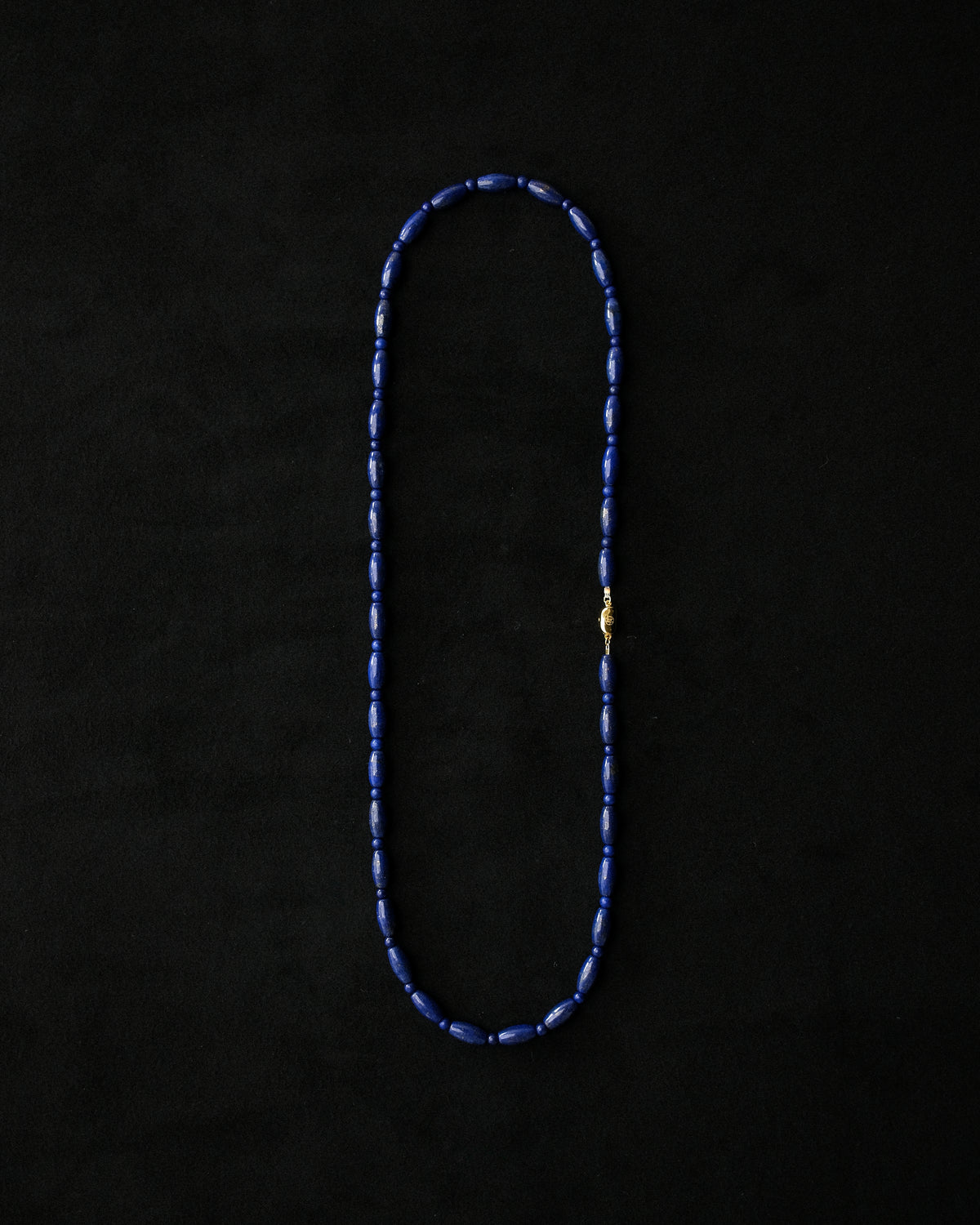 A beaded necklace made of dark blue lapis beads with a small gold clasp, laid flat on a black background.