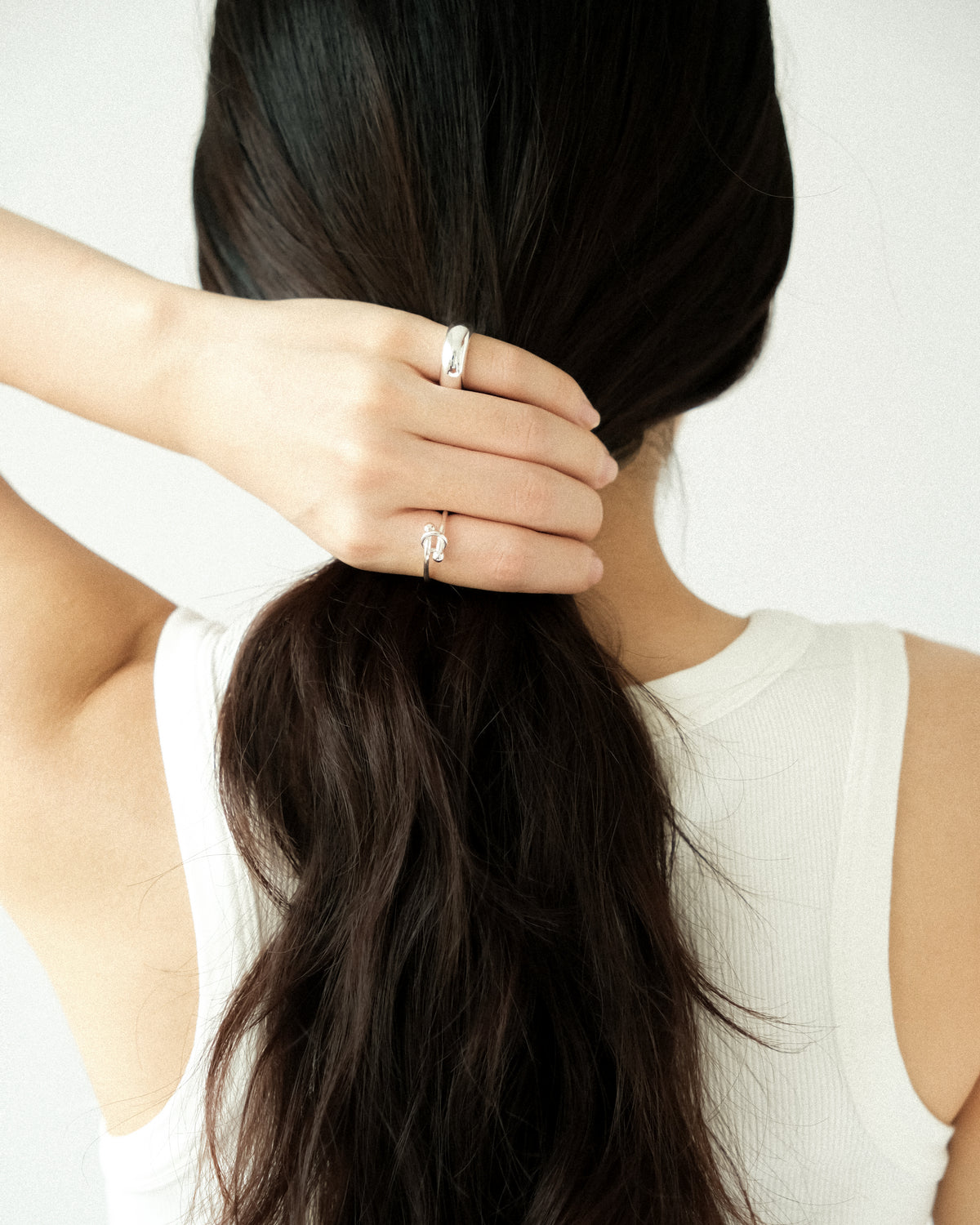 A close-up lifestyle shot of a woman with long, dark brown hair tied back in a low ponytail, with one hand holding her ponytail, wearing two silver rings: a silver knot ring on her index finger and a dome silver ring on her pinky, against a clean white background.