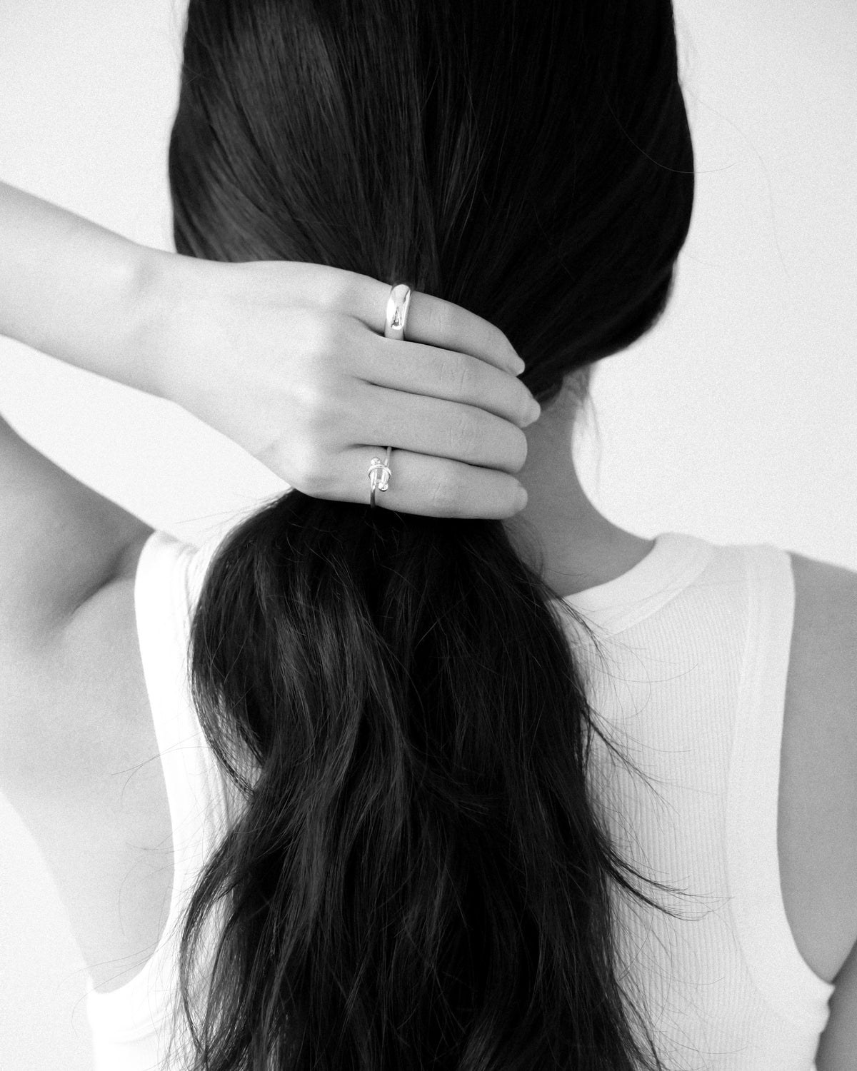 A close-up, monochrome lifestyle shot of a woman with long, dark brown hair tied back in a low ponytail, with one hand holding her ponytail, wearing two silver rings: a silver knot ring on her index finger and a dome silver ring on her pinky, against a clean white background.