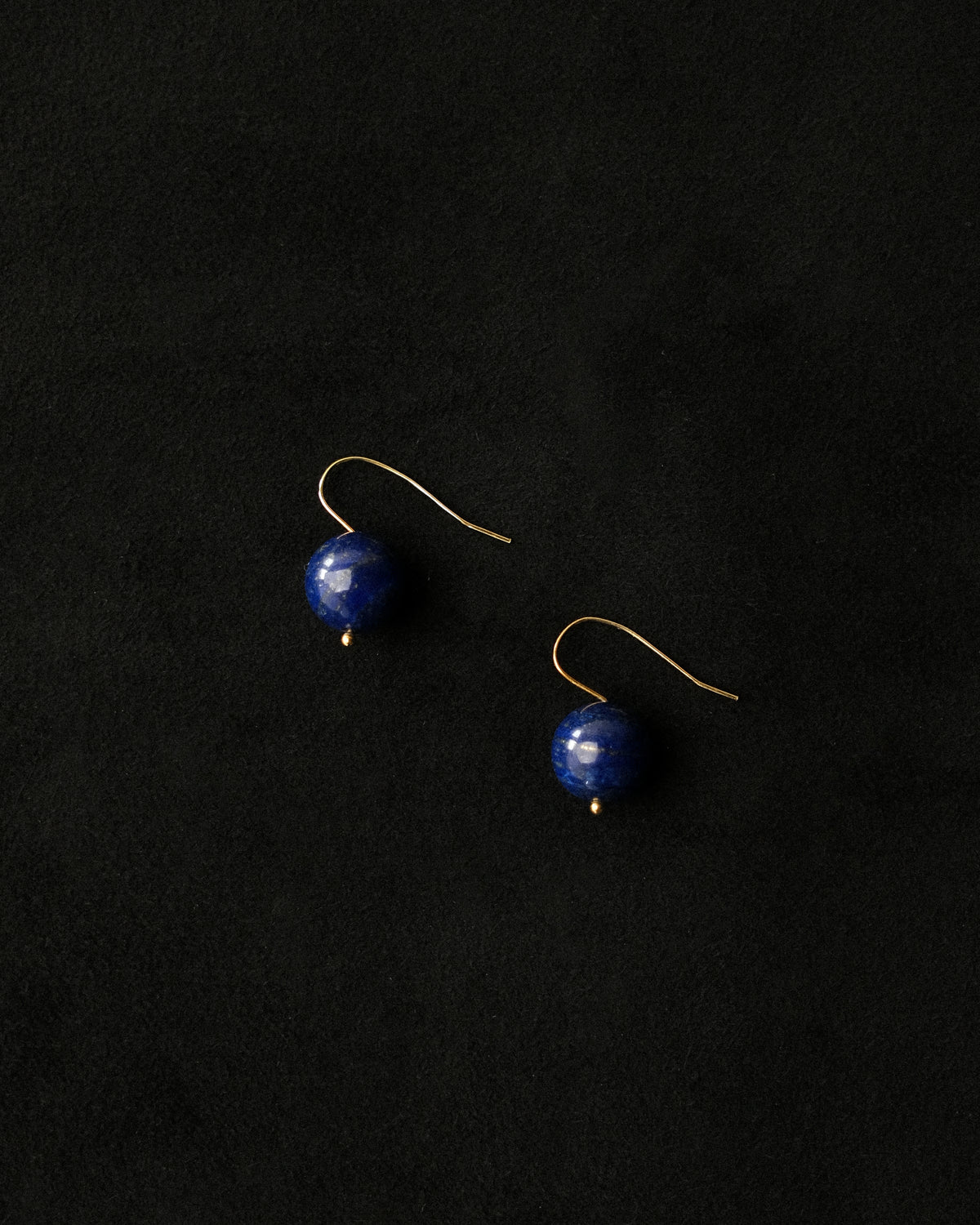 A pair of gold-tone dangle earrings with smooth, polished deep blue lapis beads, displayed on a dark textured black background.