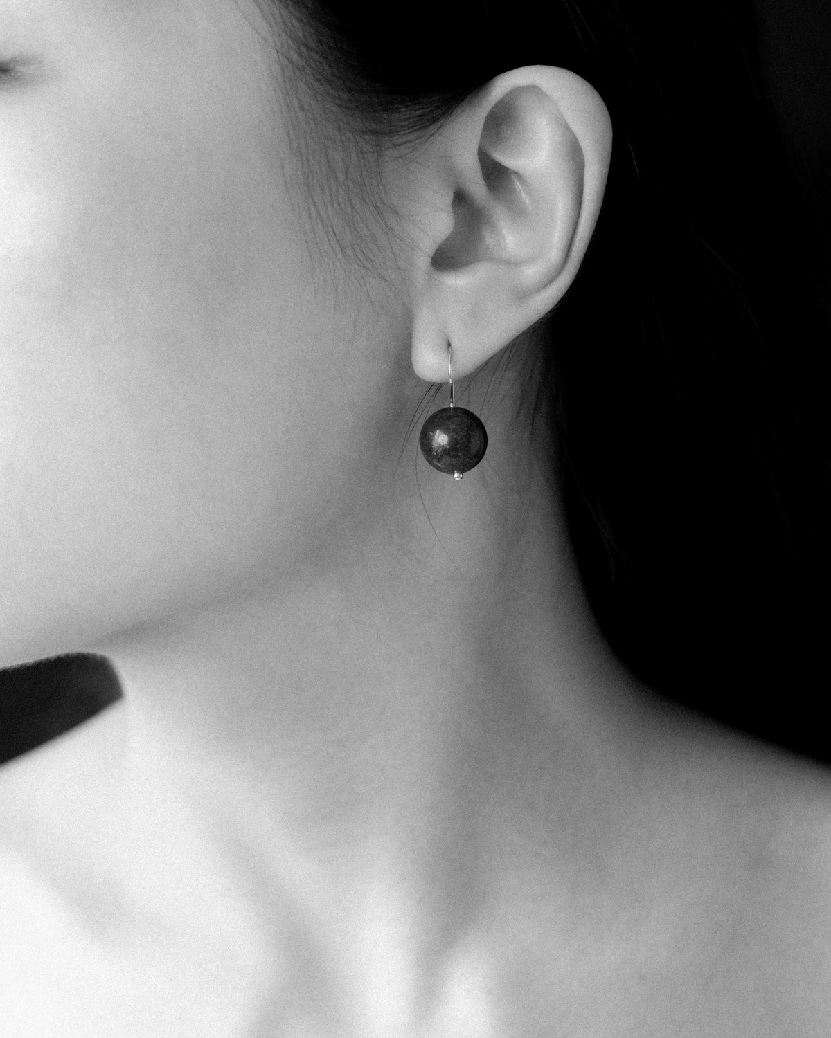 A close-up black and white photograph of a woman's ear and neck, wearing a dangling round dark blue lapis earring.