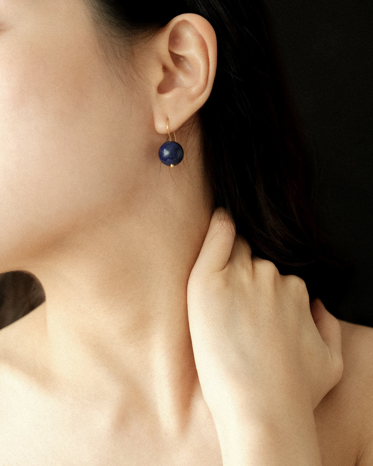 A close-up of a model wearing a single gold hoop earring with a round dark blue lapis bead drop, set against a dark background.