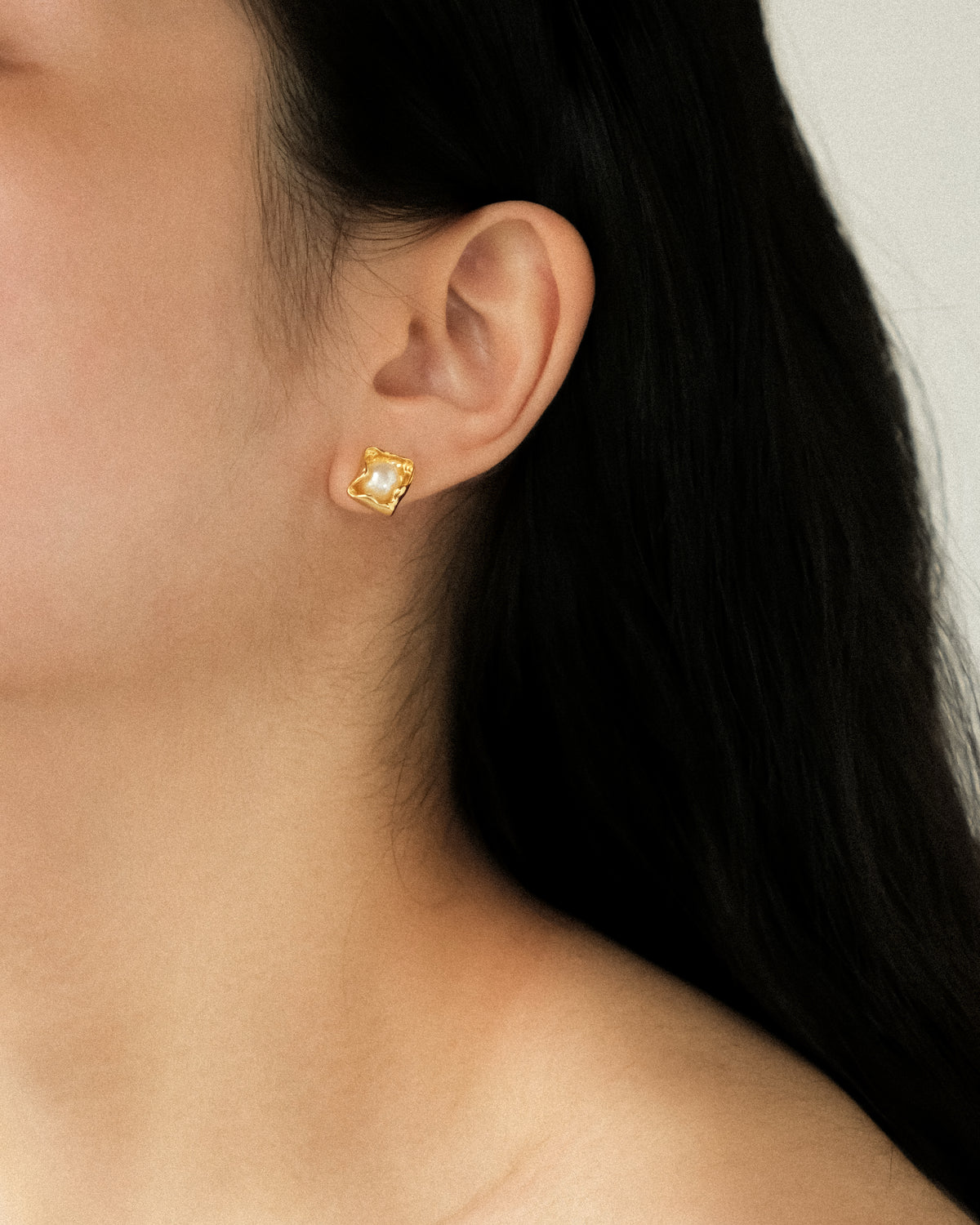 A close-up of a model’s ear wearing a small gold stud earring with a petal-shaped design featuring a central white freshwater pearl, set against a soft, neutral light background.