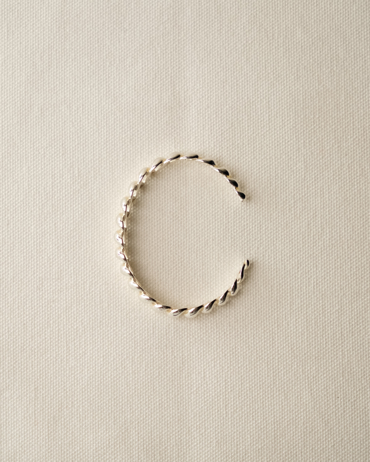 A single twisted open cuff bracelet in a sterling silver, displayed on a textured cream table cloth background.