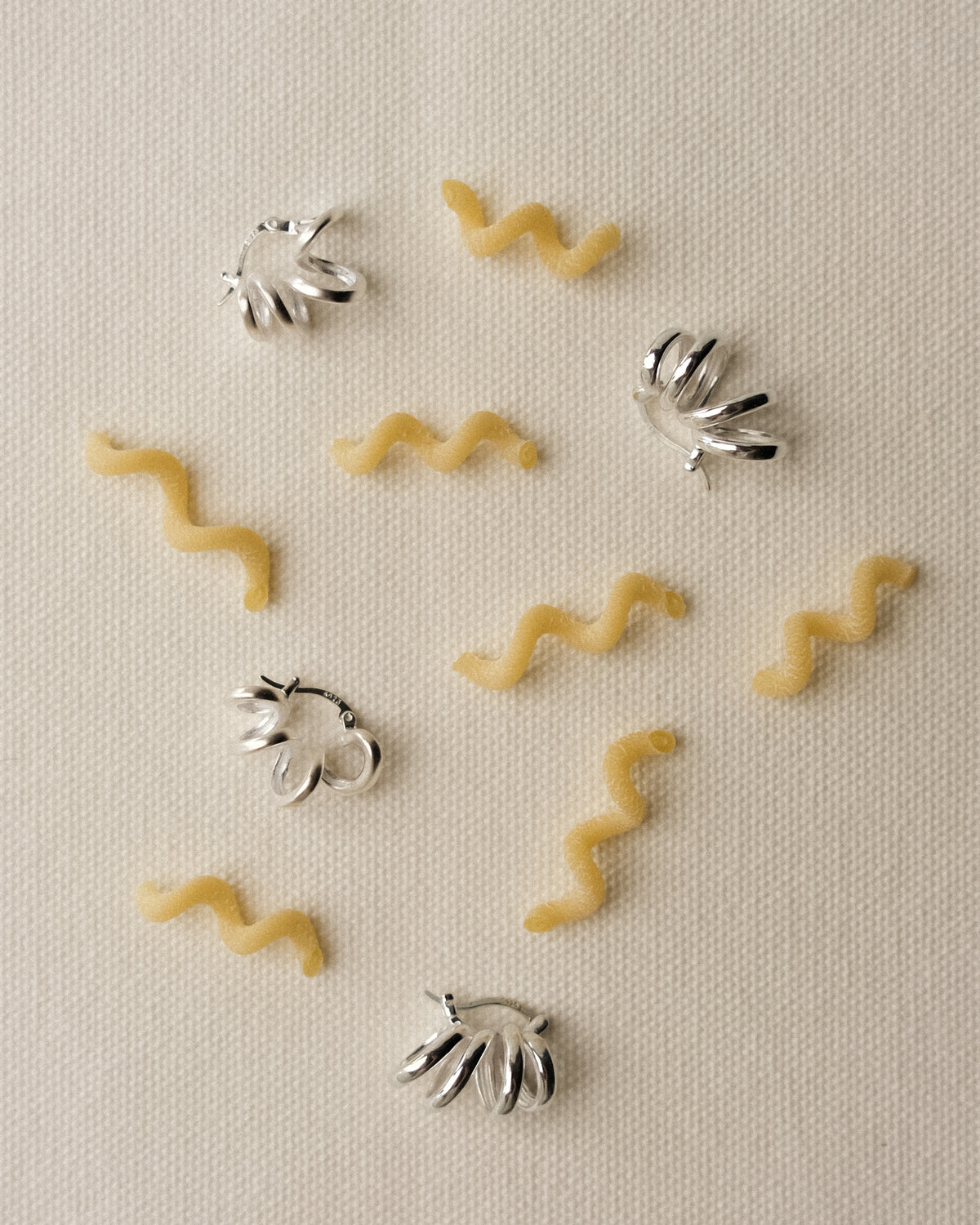 Silver sculptural hoop earrings arranged with curly fusilli bucati pasta on a textured cream table cloth.
