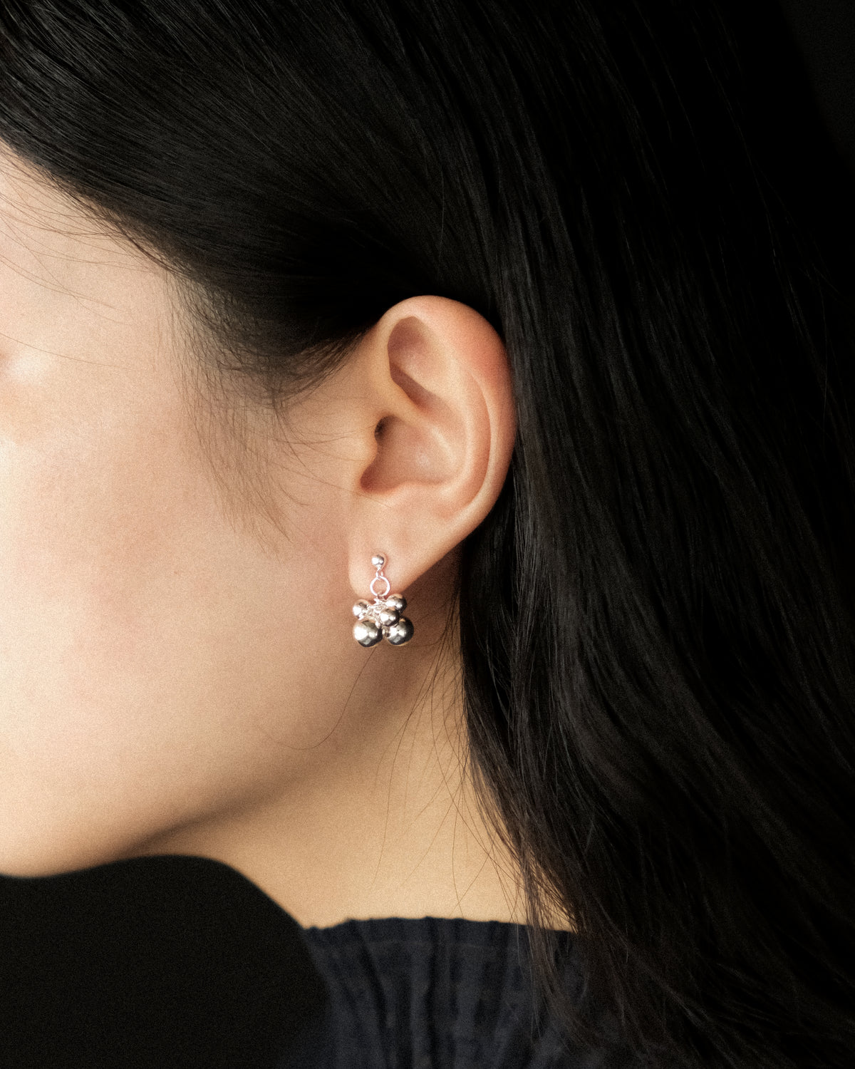 A close-up side profile of a person with dark hair wearing a small silver earring with a bunch of dangling round silver beads.