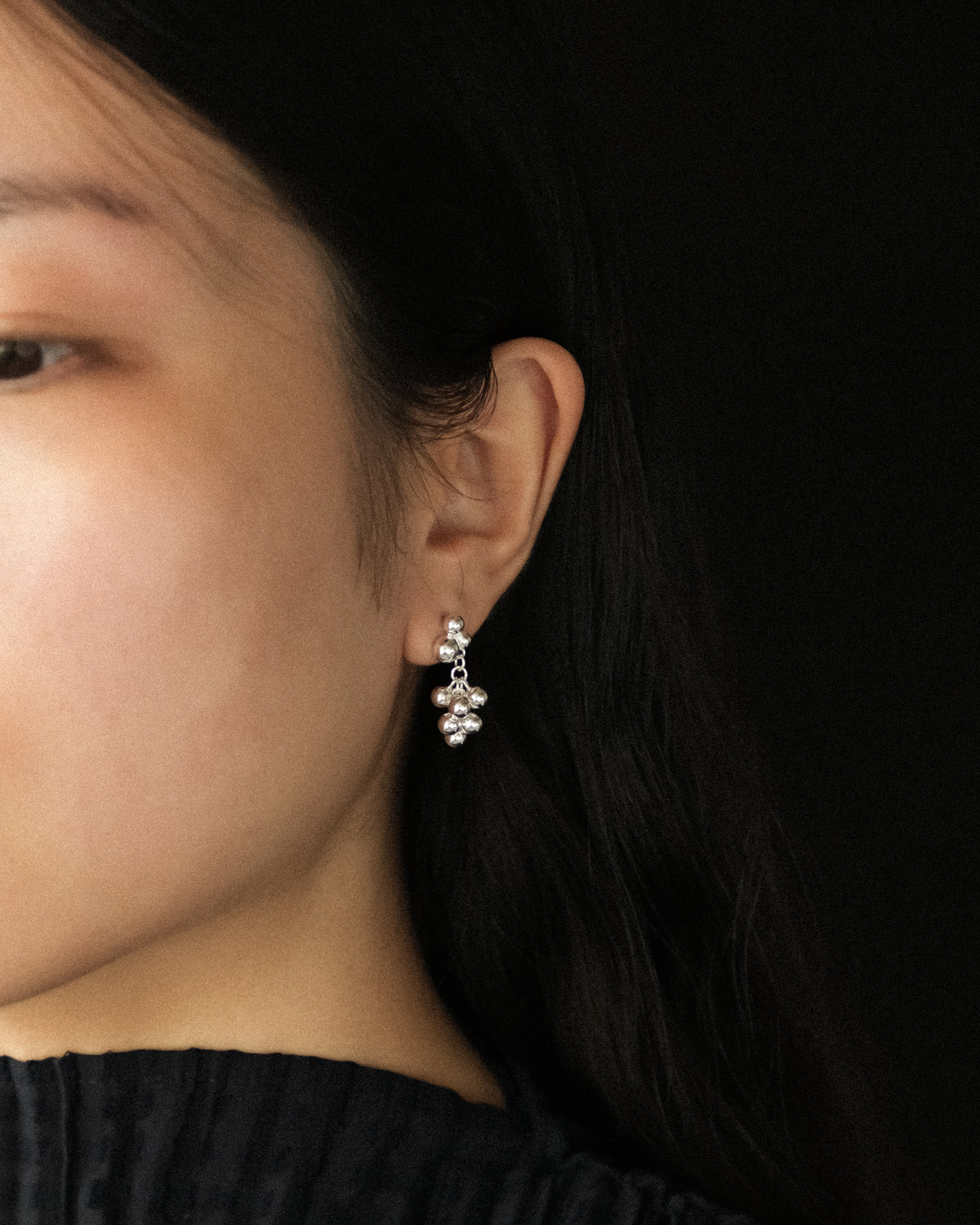 A close-up side profile of a woman wearing a small silver sphere like grape clusters dangle earring against a solid black background.