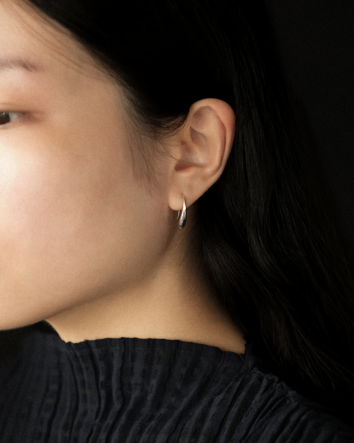 A close-up side profile of a woman with dark hair wearing a small silver crescent-shaped hoop earring, set against a dark background.